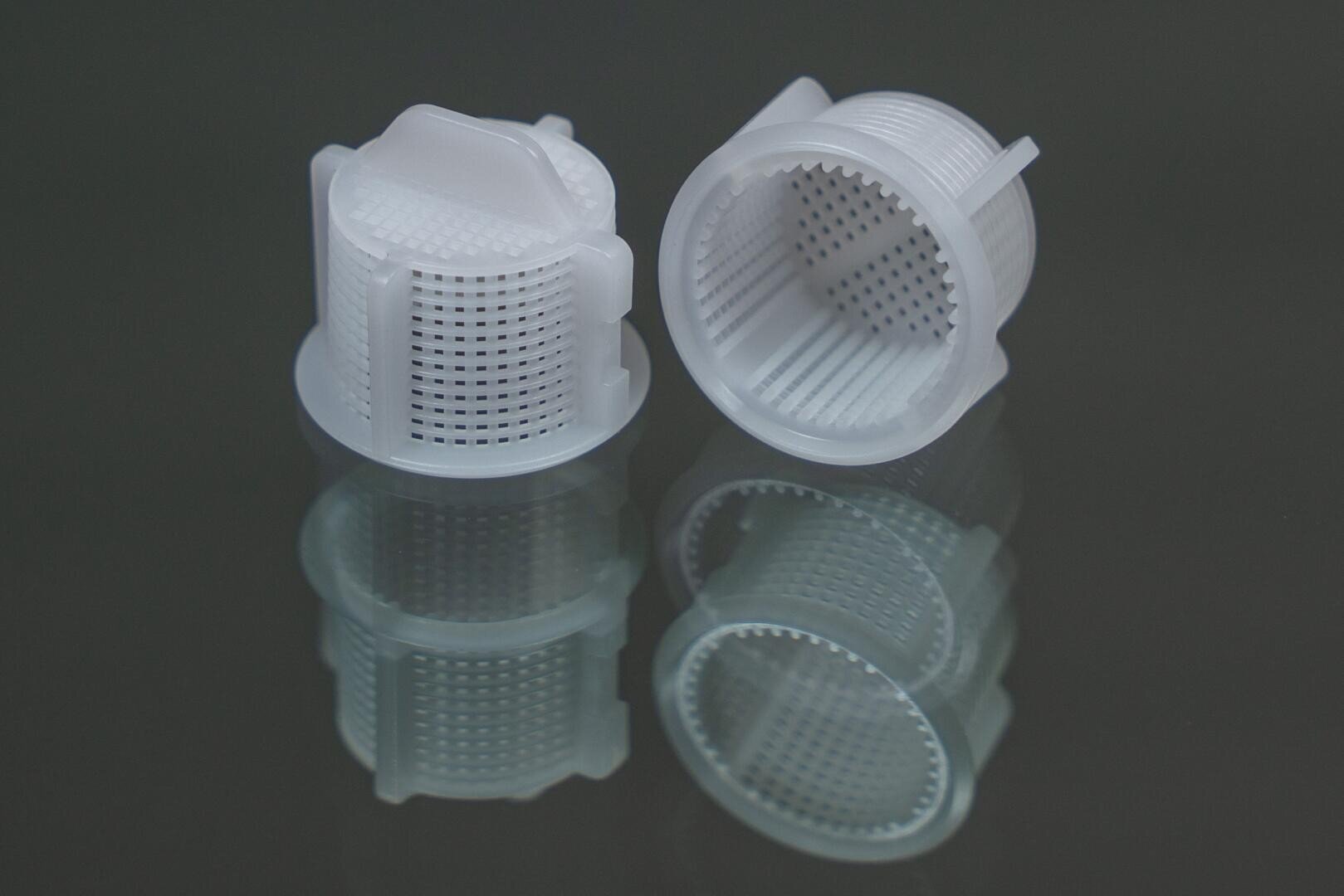 Auf einer spiegelnden, dunklen Fläche sind zwei weiße, zylindrische Kunststofffilter mit netzartigen Perforationen abgebildet. Ein Filter steht aufrecht, während der andere auf der Seite liegt und sein hohles Inneres zeigt.