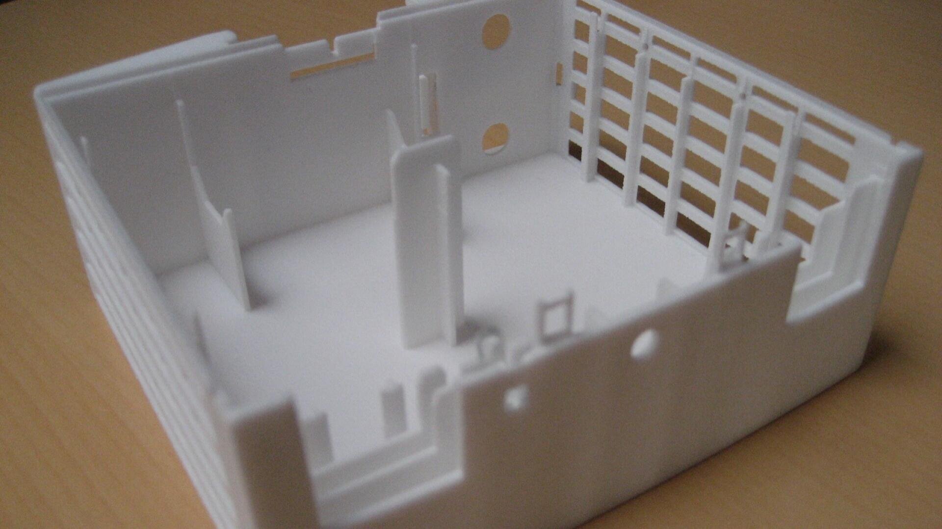 Ein weißes, rechteckiges Kunststoffgehäuse mit Ausschnitten, Schlitzen und inneren Halterungen, wahrscheinlich ein Gehäuse für elektronische Bauteile, steht auf einer hellen Holzoberfläche.