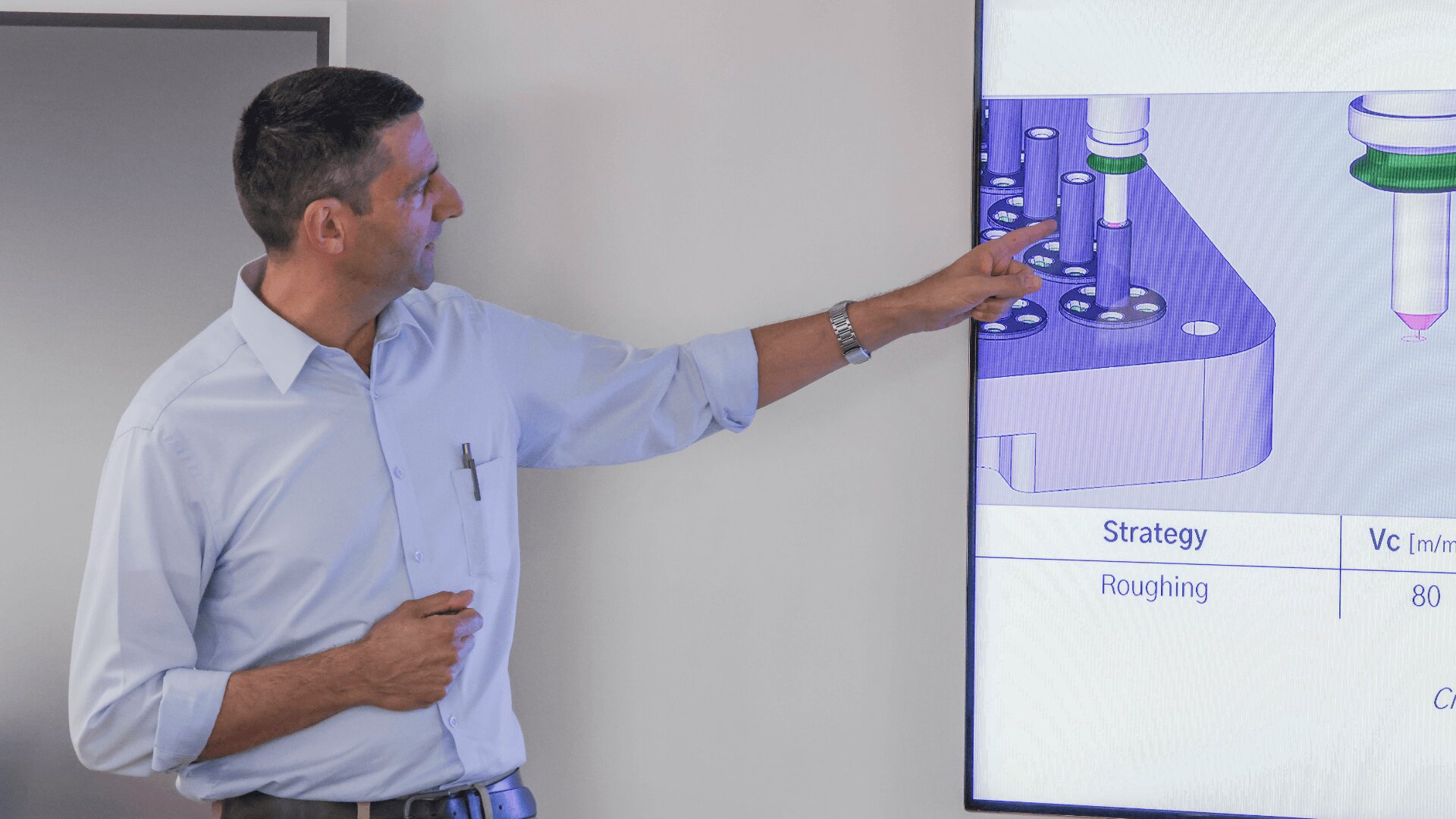 Ein Mann in einem hellblauen Hemd zeigt auf ein technisches Diagramm auf einem großen Bildschirm und erklärt Bearbeitungsstrategien mit den Bezeichnungen „Strategie“, „Schruppen“ und „Vc“. Er steht neben dem Bildschirm in einem Klassenzimmer oder Büro.