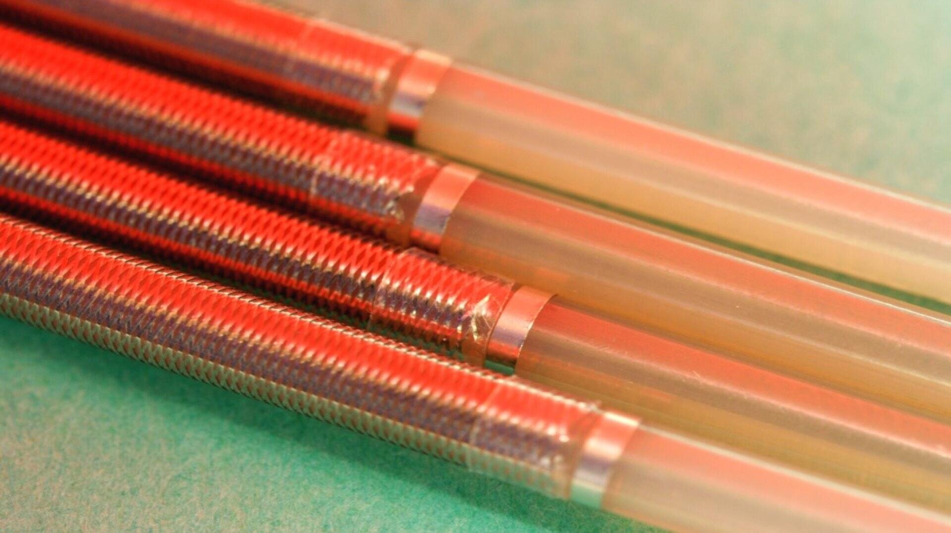 Nahaufnahme von vier metallischen Druckbleistiftspitzen und durchscheinenden Schäften, die parallel auf einer grünen Oberfläche angeordnet und mit rotem und orangefarbenem Licht beleuchtet sind.