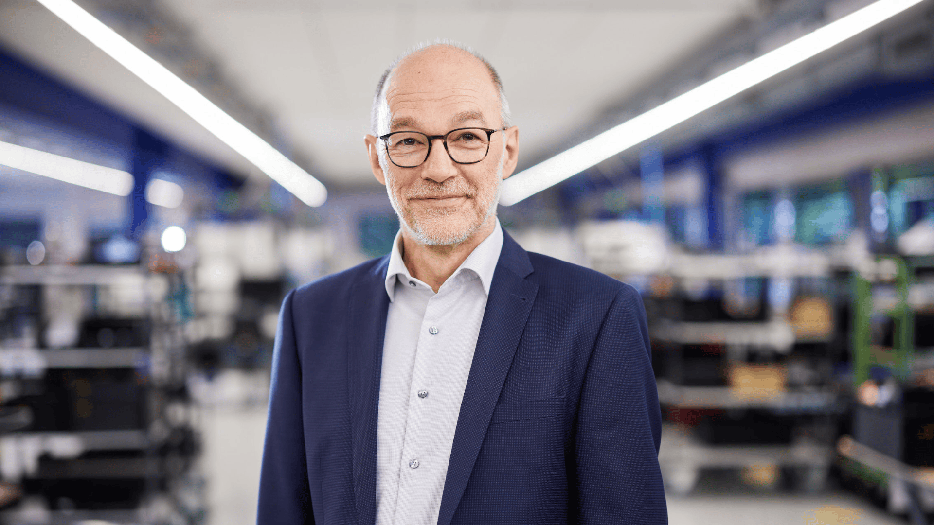 Ein Mann mittleren Alters mit Brille, kurzem grauem Bart und Glatze, der einen blauen Anzug und ein helles Hemd trägt, steht in einem hellen, modernen Büro oder Labor, schaut in die Kamera und lächelt sanft.