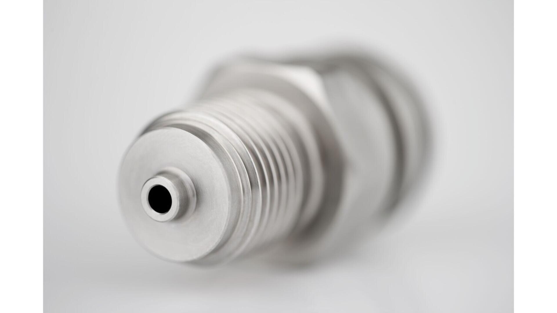 Close-up view of a metallic spark plug with a threaded body, photographed against a white background. The focus is on the tip, highlighting the detailed grooves and central hole.