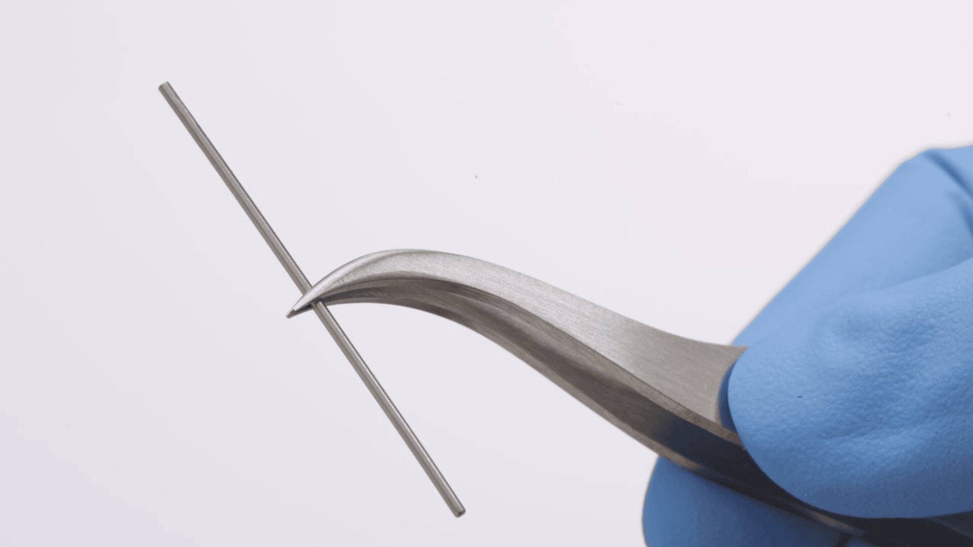 Eine behandschuhte Hand hält eine gebogene Metallpinzette und greift horizontal einen dünnen Metallstab vor einem hellen Hintergrund.