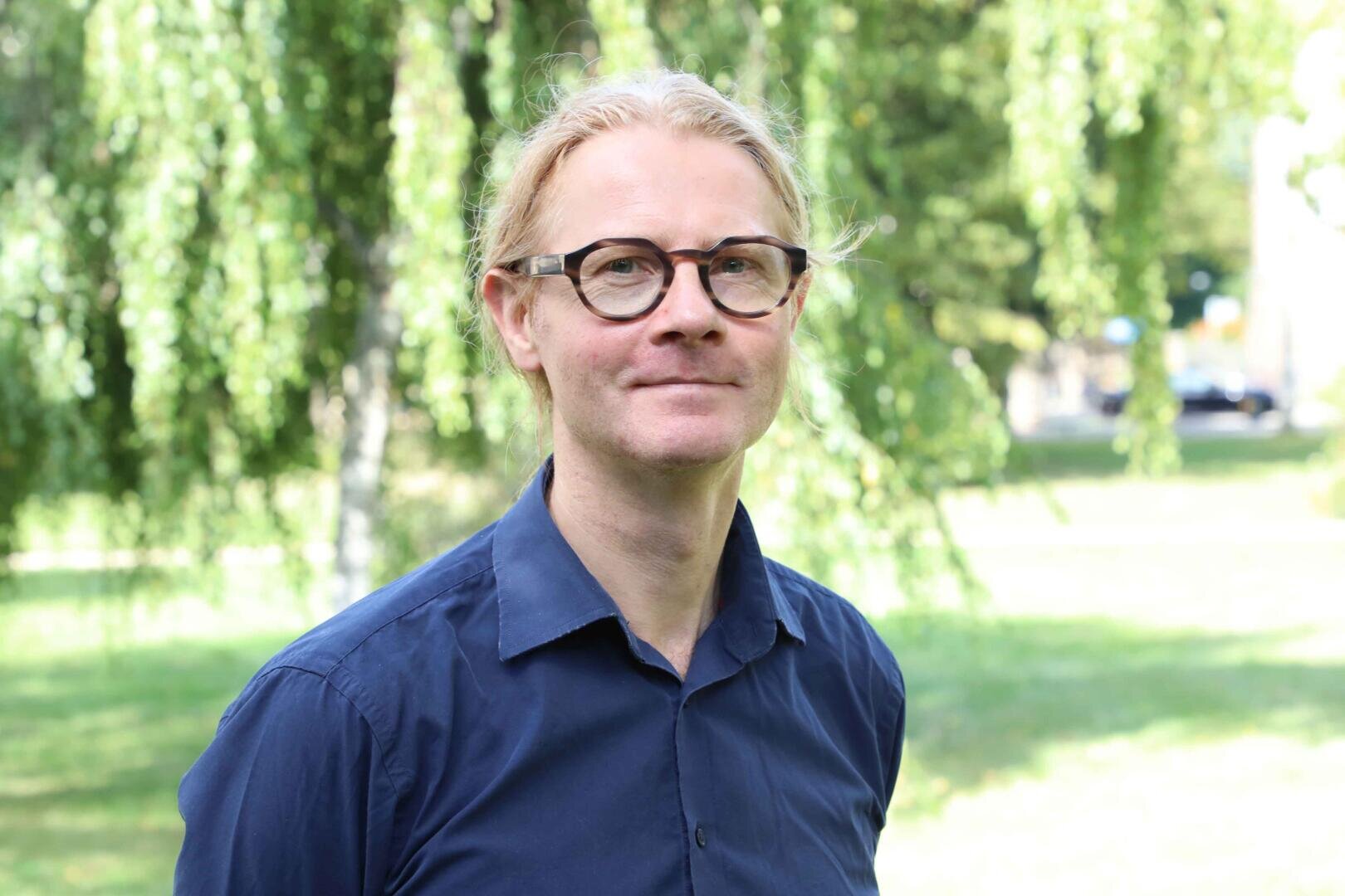 Dr. Stephan Gilbert mit blondem Haar und Brille, die ein marineblaues Hemd trägt, steht im Freien in einem Park mit grünen Bäumen und Gras im Hintergrund.