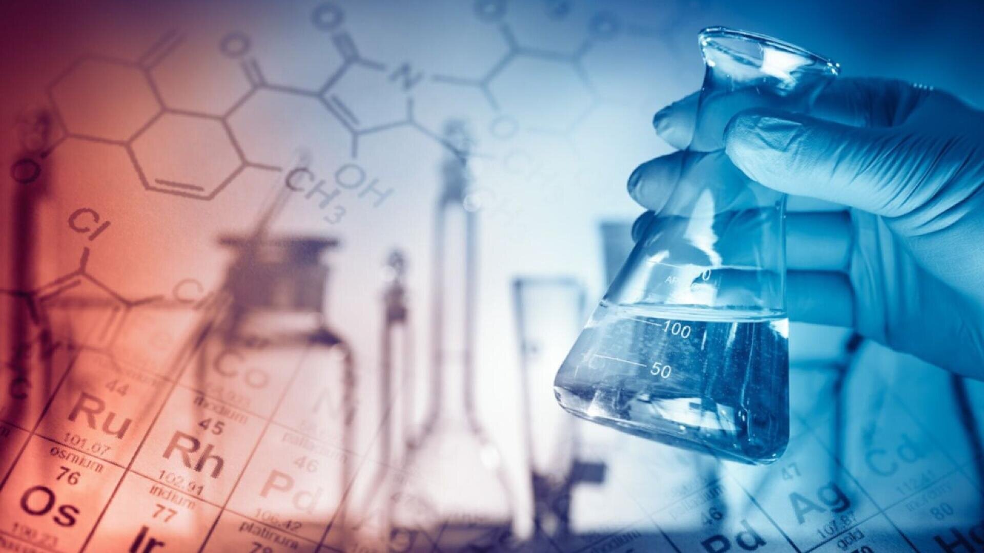 A gloved hand holds a glass flask with liquid in a laboratory setting, with blurred chemical formulas, lab glassware, and a periodic table in the blue and red-tinted background.