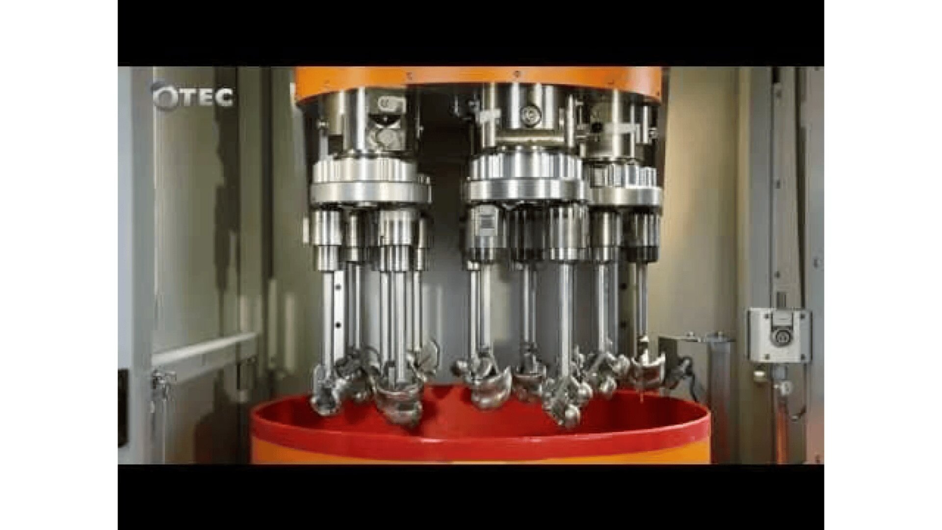 Nahaufnahme einer industriellen Poliermaschine mit mehreren rotierenden Spindeln und Metallteilen, die über einem roten Becken aufgehängt ist, mit dem OTEC-Logo in der oberen linken Ecke.