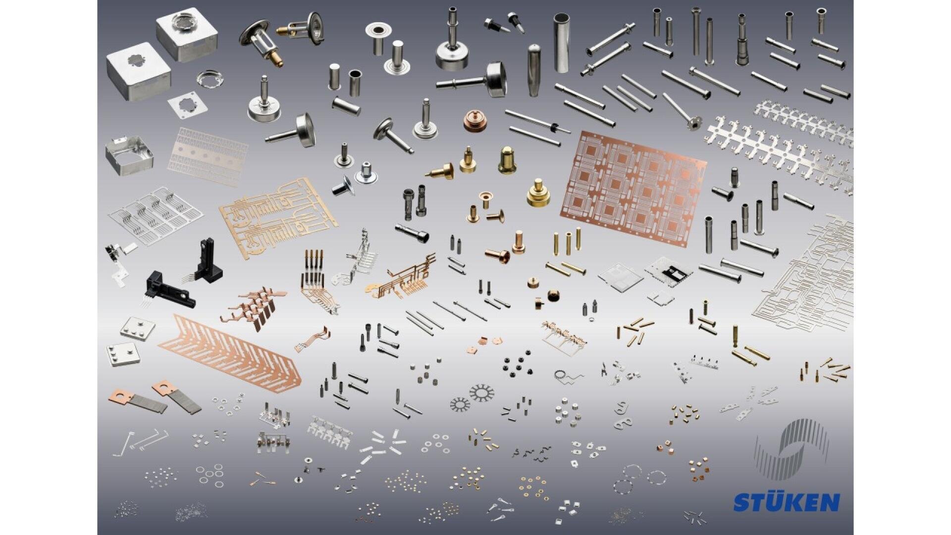 A wide assortment of precision metal parts and components, including pins, screws, plates, and springs, arranged on a gradient gray background with the STÜKEN logo in the lower right corner.