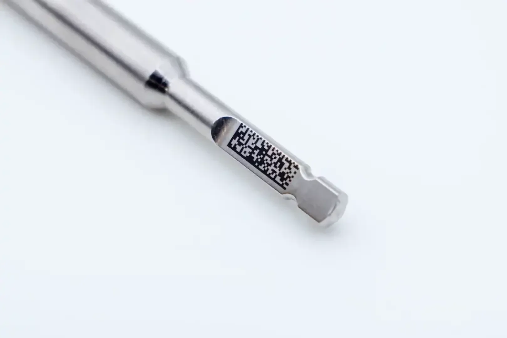 Close-up of a metallic tool or drill, with a small black and white QR code or data matrix engraved on its shank, on a plain light background.