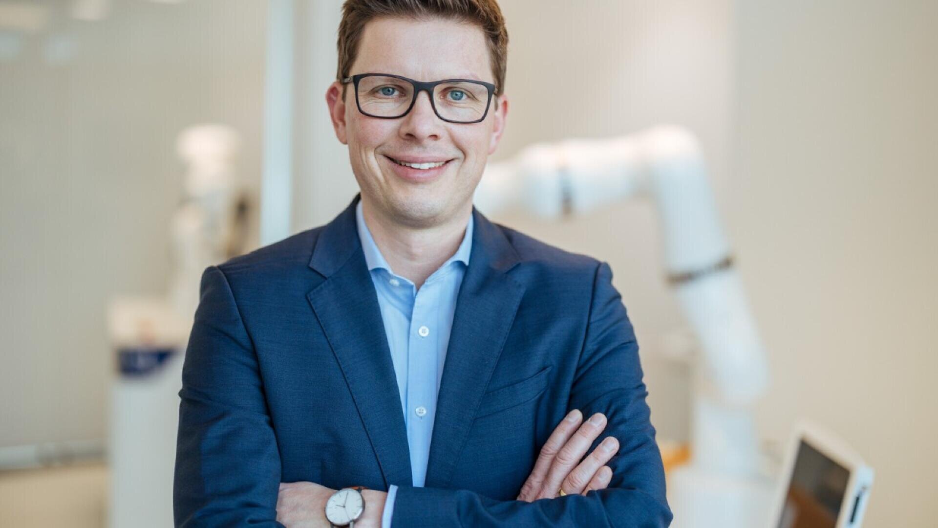 A man wearing glasses and a blue suit stands with arms crossed, smiling in a bright, modern setting with blurred white robotic equipment in the background.