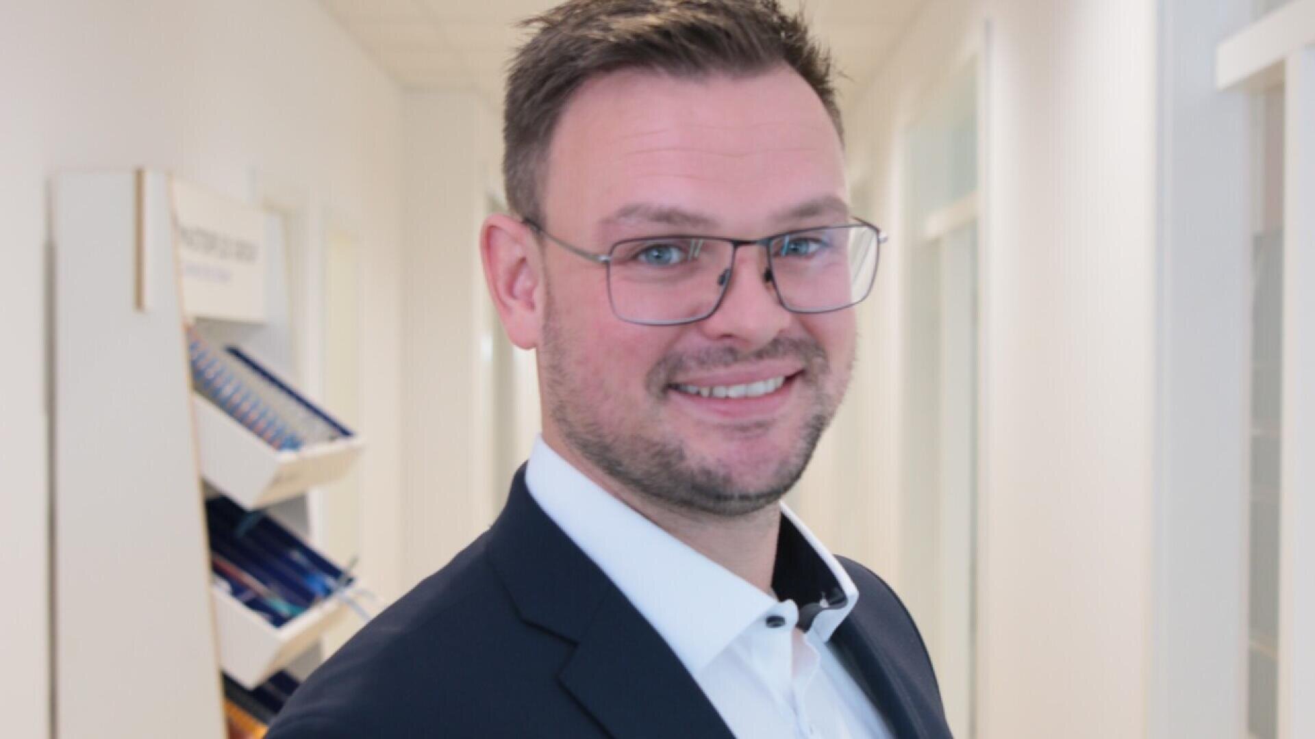 A man with short brown hair, glasses, and a beard is smiling while wearing a dark suit and white shirt. He stands in a bright hallway with shelves of folders in the background.
