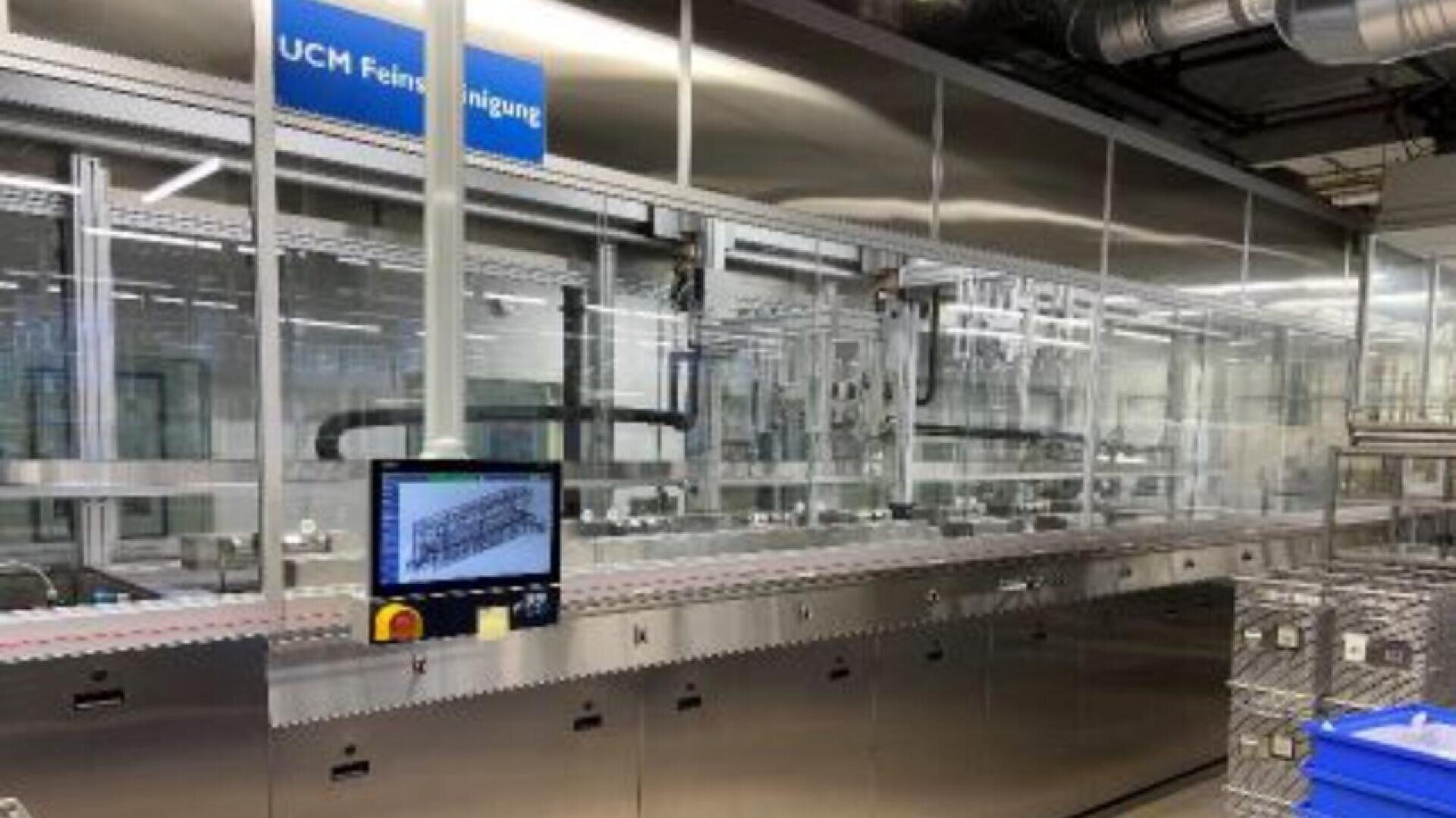 A modern industrial cleaning machine with a touchscreen control panel in a bright, reflective factory setting. A blue sign above reads UCM Feinstreinigung. Metal baskets are visible on the right side.