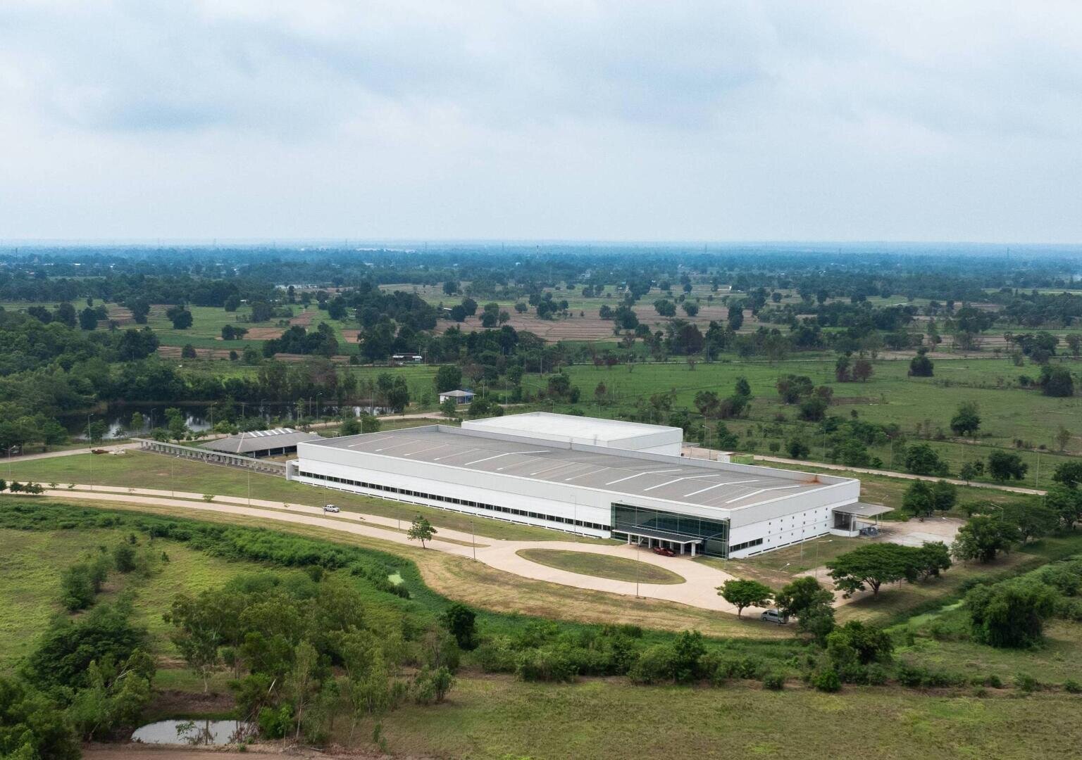 Ein großes, modernes, weißes Industriegebäude mit Flachdach ist umgeben von grünen Feldern und Bäumen unter einem wolkenverhangenen Himmel, mit einer Straße, die zum Eingang führt, und einer fernen Landschaft im Hintergrund.