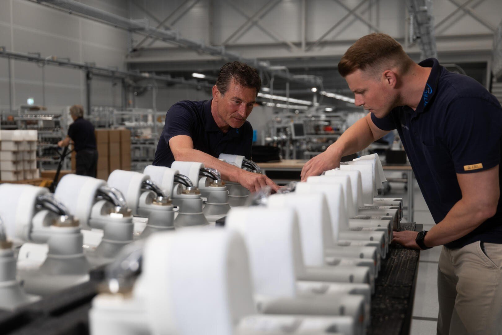 Two men inspect and assemble white machine parts lined up on a workbench in a factory; industrial plants and shelves can be seen in the background.