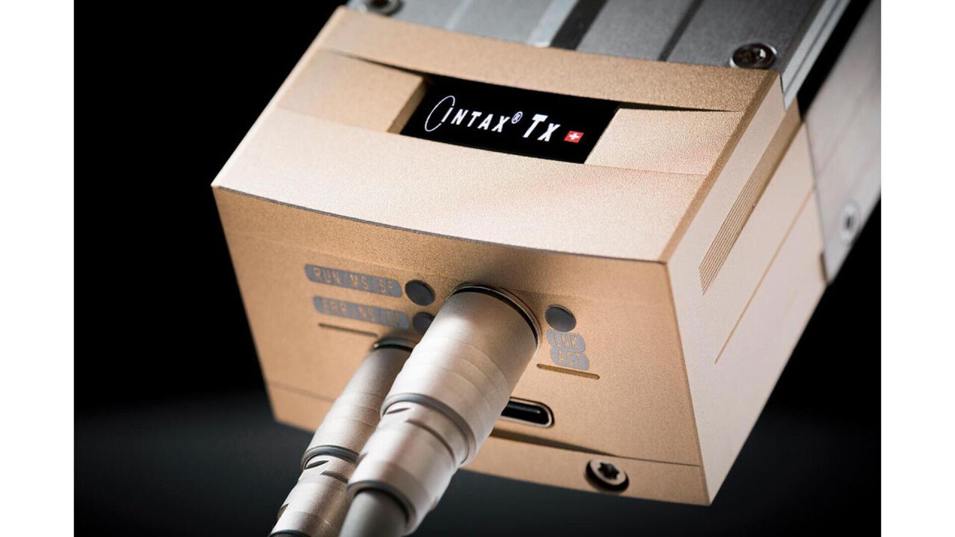Close-up of a gold-colored electronic device labeled OMTAX Tx, with two metal cables plugged into the front ports and a USB-C port visible between them on a black background.