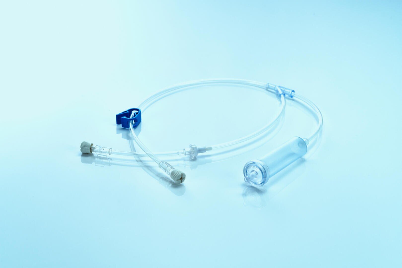 A sterile medical IV infusion set with clear tubing, a blue clamp, connectors and a drip chamber is placed on a light blue reflective surface.
