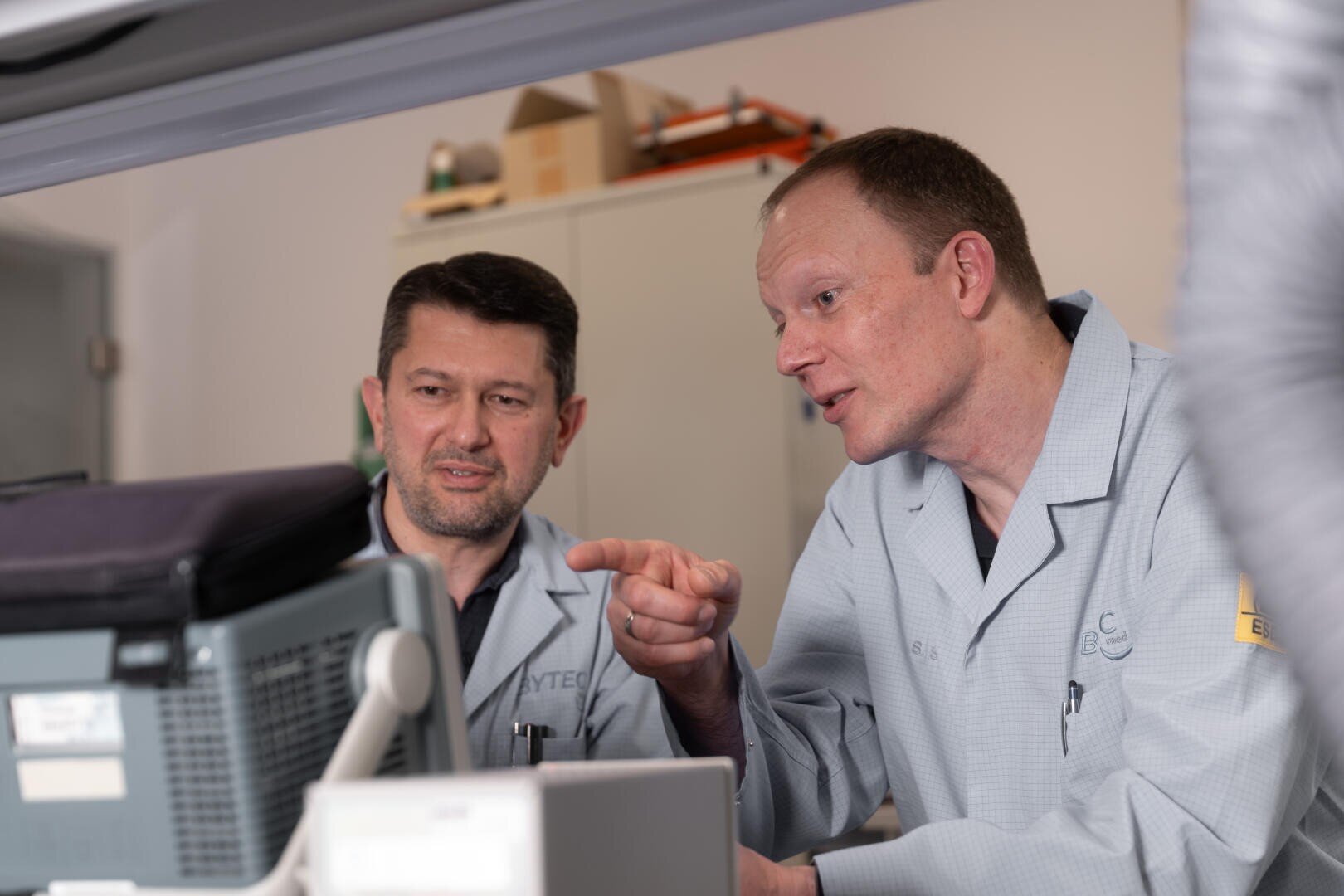 Two men in lab coats are looking at a computer monitor. One man points at the screen while explaining something to the other, who listens attentively. They appear to be working in a laboratory or technical environment.