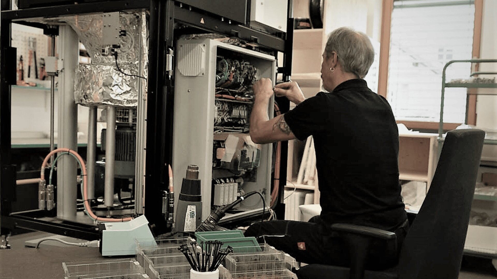 Ein Mann sitzt auf einem Stuhl und repariert oder montiert eine Industriemaschine oder ein großes elektronisches Gerät, umgeben von Werkzeugen und Teilen in einer Werkstattumgebung.