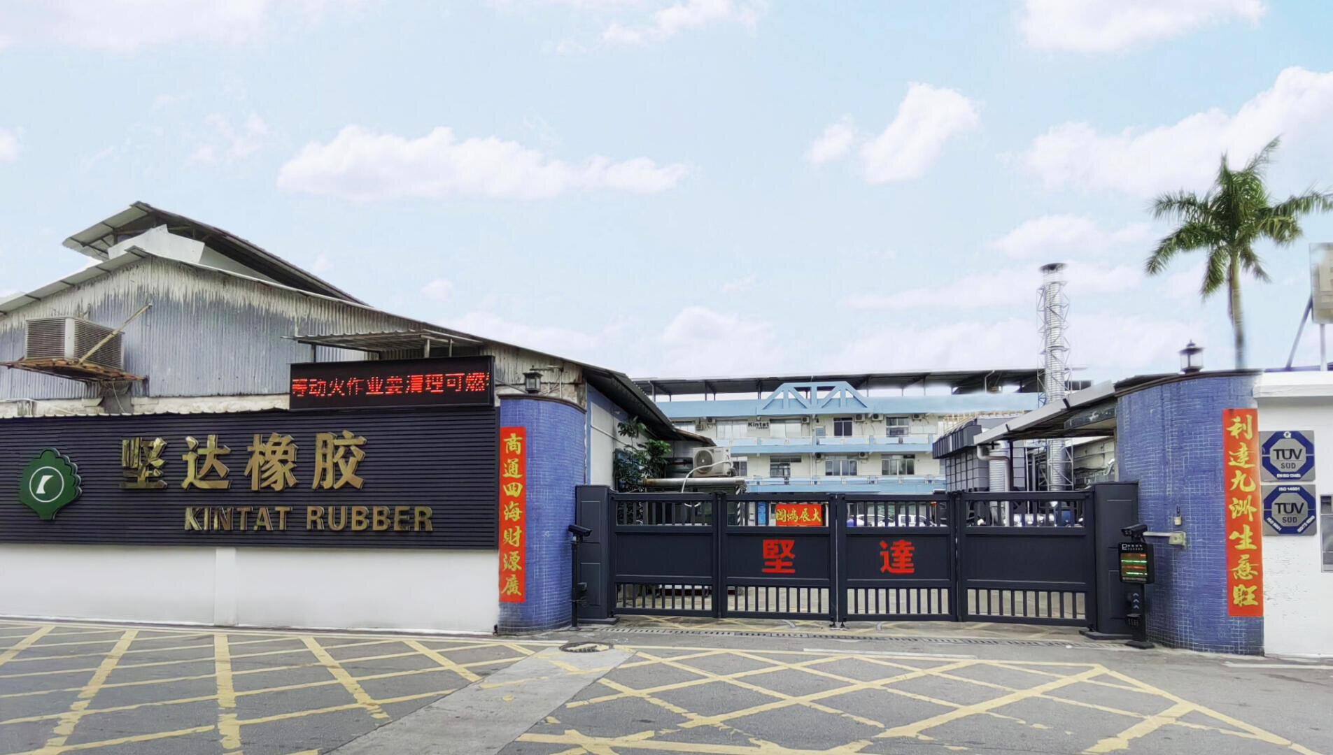 The entrance gate of Kintat Rubber factory with signs in English and Chinese, blue walls, and a palm tree to the right. Industrial buildings are visible in the background under a partly cloudy sky.