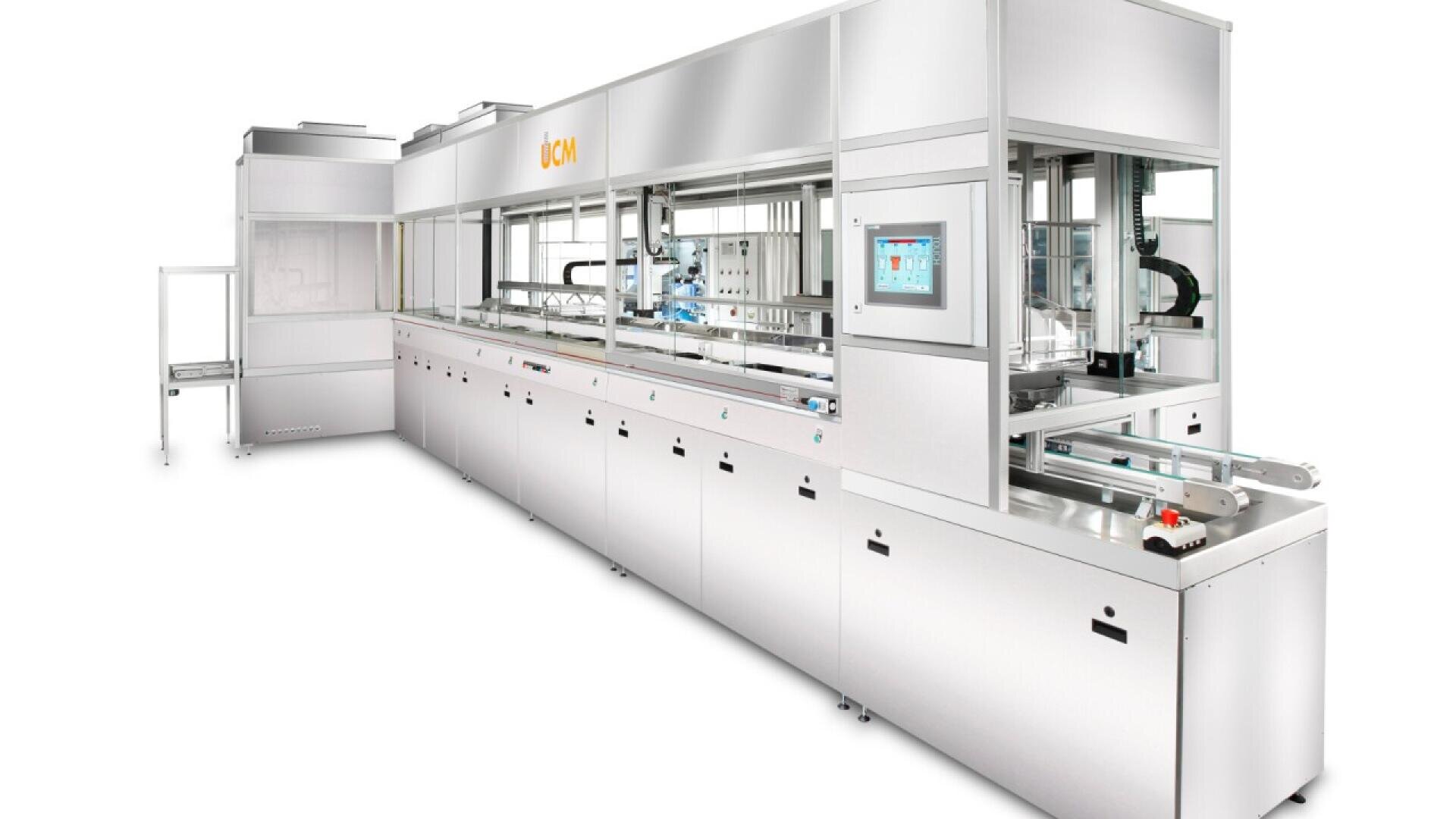 A large, modern industrial cleaning machine with a stainless steel finish, transparent panels, conveyor system, and a touchscreen control panel. The machine is clean and situated in a bright, white room.