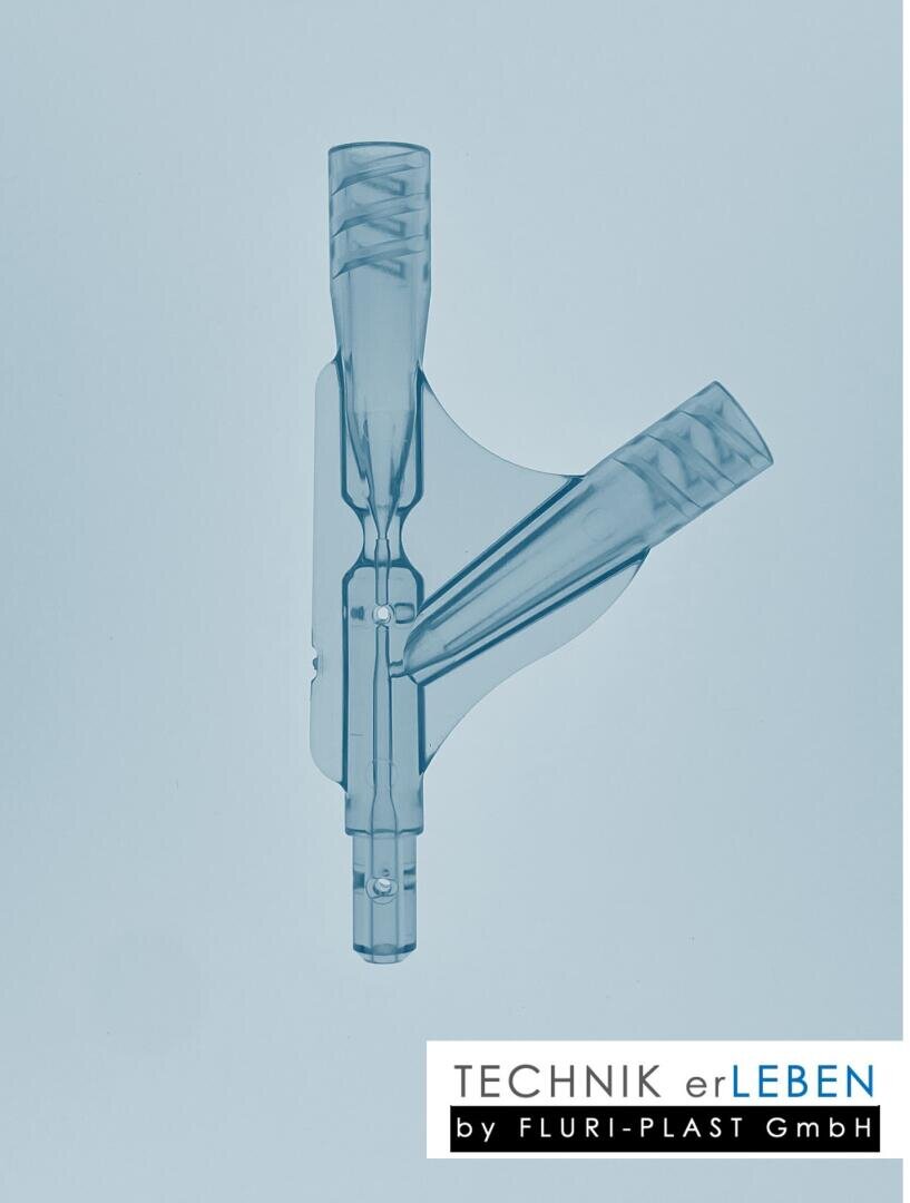 A translucent blue plastic part with two angled tubes attached to a centerpiece, shown against a light background. The TECHNIK erLEBEN by FLURI-PLAST GmbH logo is shown in the bottom right-hand corner.
