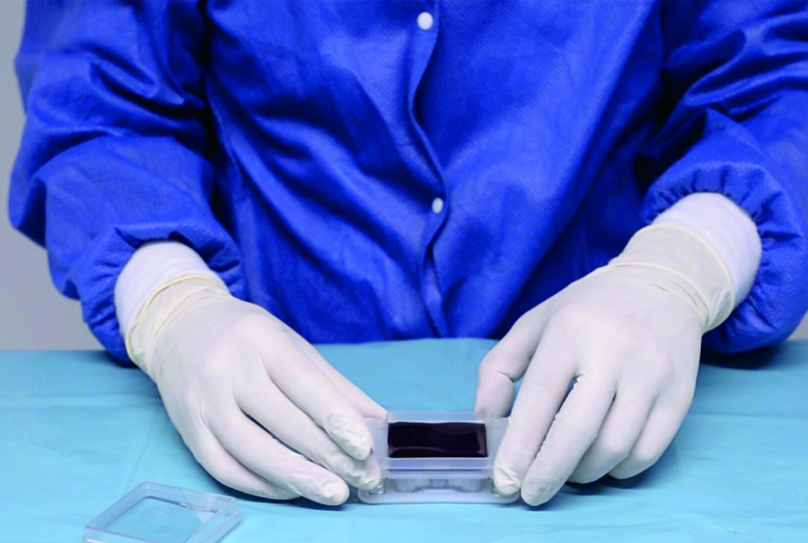 Eine Person in einem blauen Laborkittel und mit weißen Handschuhen hantiert mit einem kleinen durchsichtigen Behälter, in dem sich ein dunkler Gegenstand befindet, auf einer hellblauen Fläche, die an ein Labor oder eine sterile Umgebung erinnert.