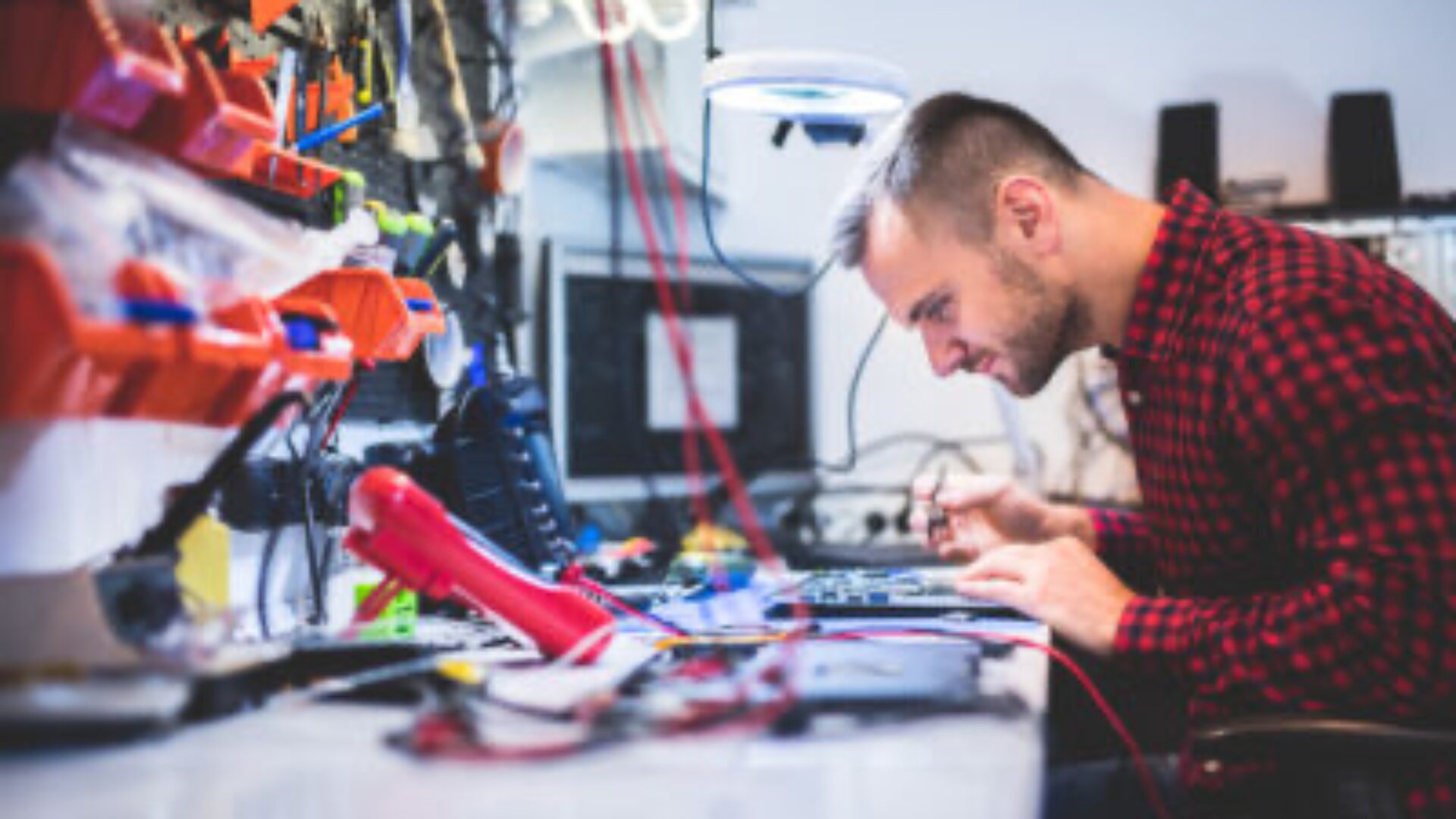 Ein Mann in einem rotkarierten Hemd sitzt an einer unübersichtlichen Werkbank und repariert Elektronik mit einem Lötkolben und verschiedenen Werkzeugen, umgeben von Kabeln und elektronischen Bauteilen.