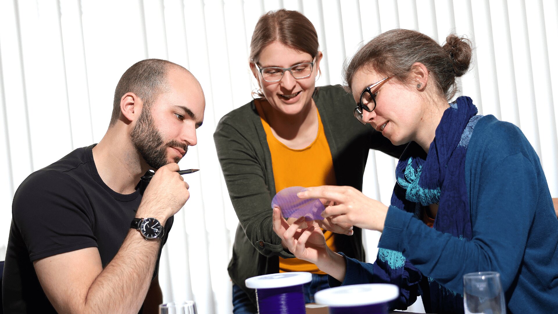 Drei Personen sitzen an einem Tisch und betrachten einen durchscheinenden violetten Gegenstand. Eine Person hält den Gegenstand, eine andere betrachtet ihn genau, und die dritte steht lächelnd und beobachtend dahinter. Helles Licht fällt durch die Jalousien hinter ihnen.