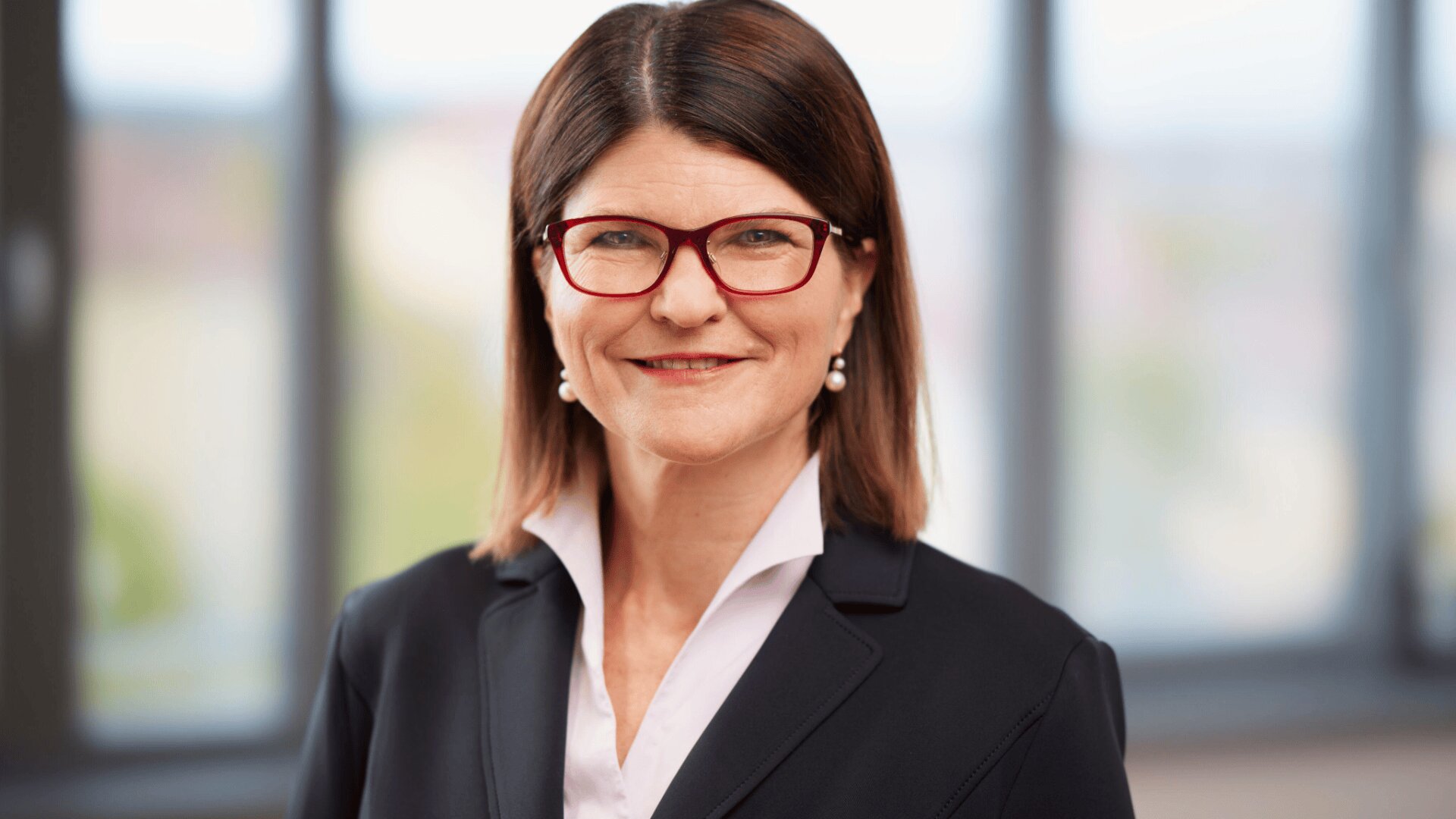 Eine Frau mit glattem braunem Haar, roter Brille, Perlenohrringen, weißer Bluse und dunklem Blazer lächelt in die Kamera. Der Hintergrund ist unscharf und lässt auf eine Inneneinrichtung mit großen Fenstern schließen.