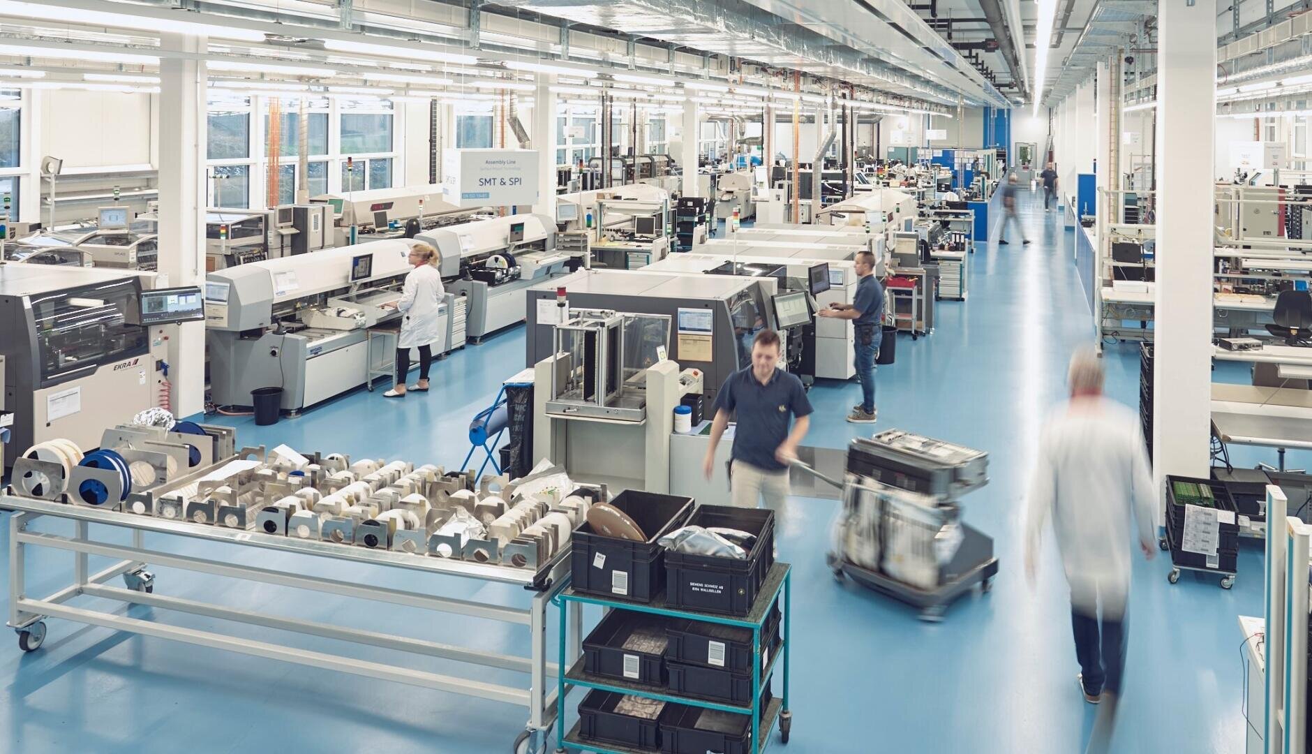 A spacious, brightly lit factory hall with a blue floor, various industrial machines and several people in lab coats and work clothes operating equipment and moving trolleys.