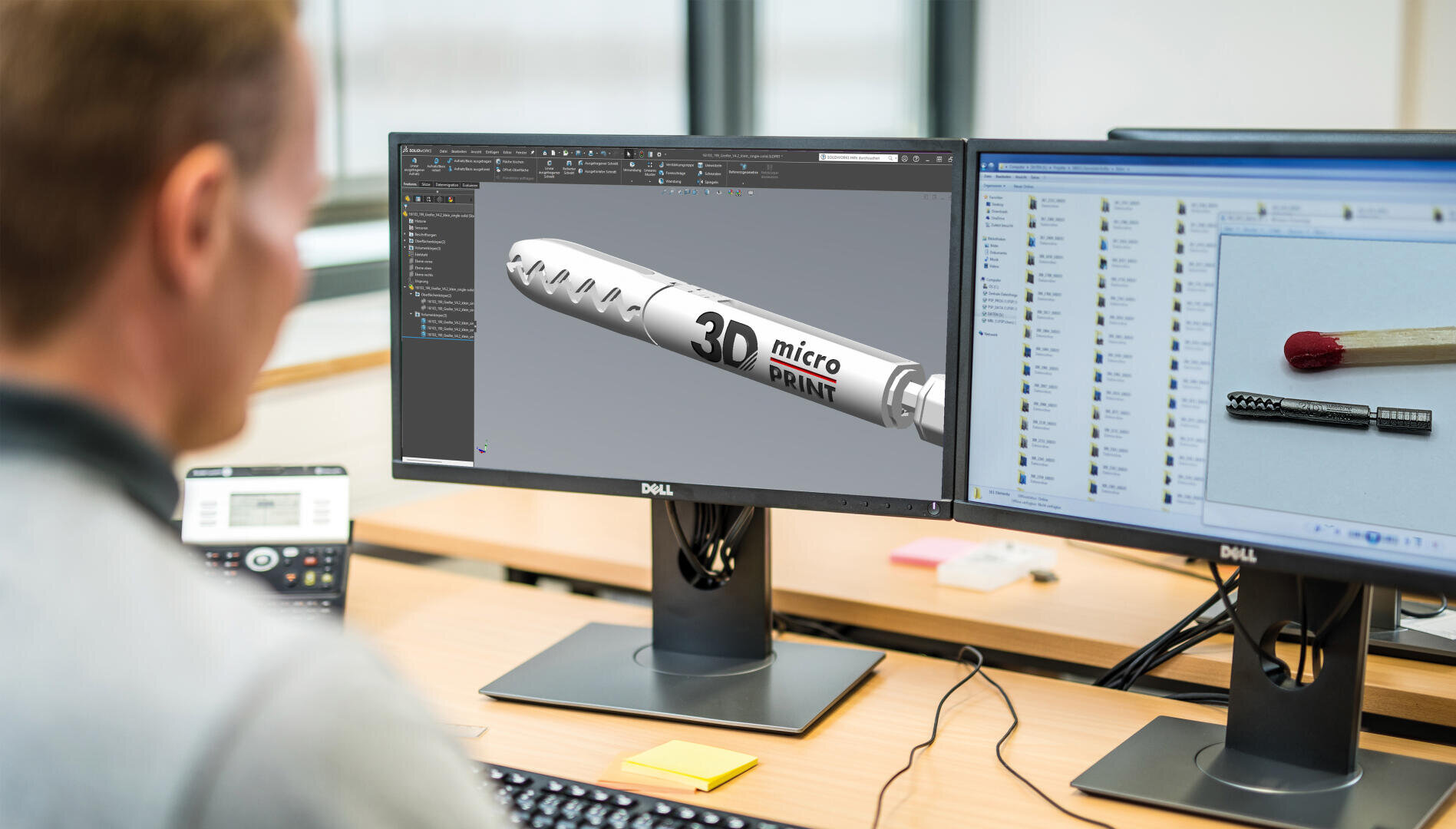 Eine Person sitzt an einem Schreibtisch mit zwei Monitoren; auf dem einen ist ein 3D-Modell mit der Aufschrift 3D-Mikrodruck zu sehen, auf dem anderen ein Foto eines kleinen Metallobjekts neben einem Streichholz als Maßstab.