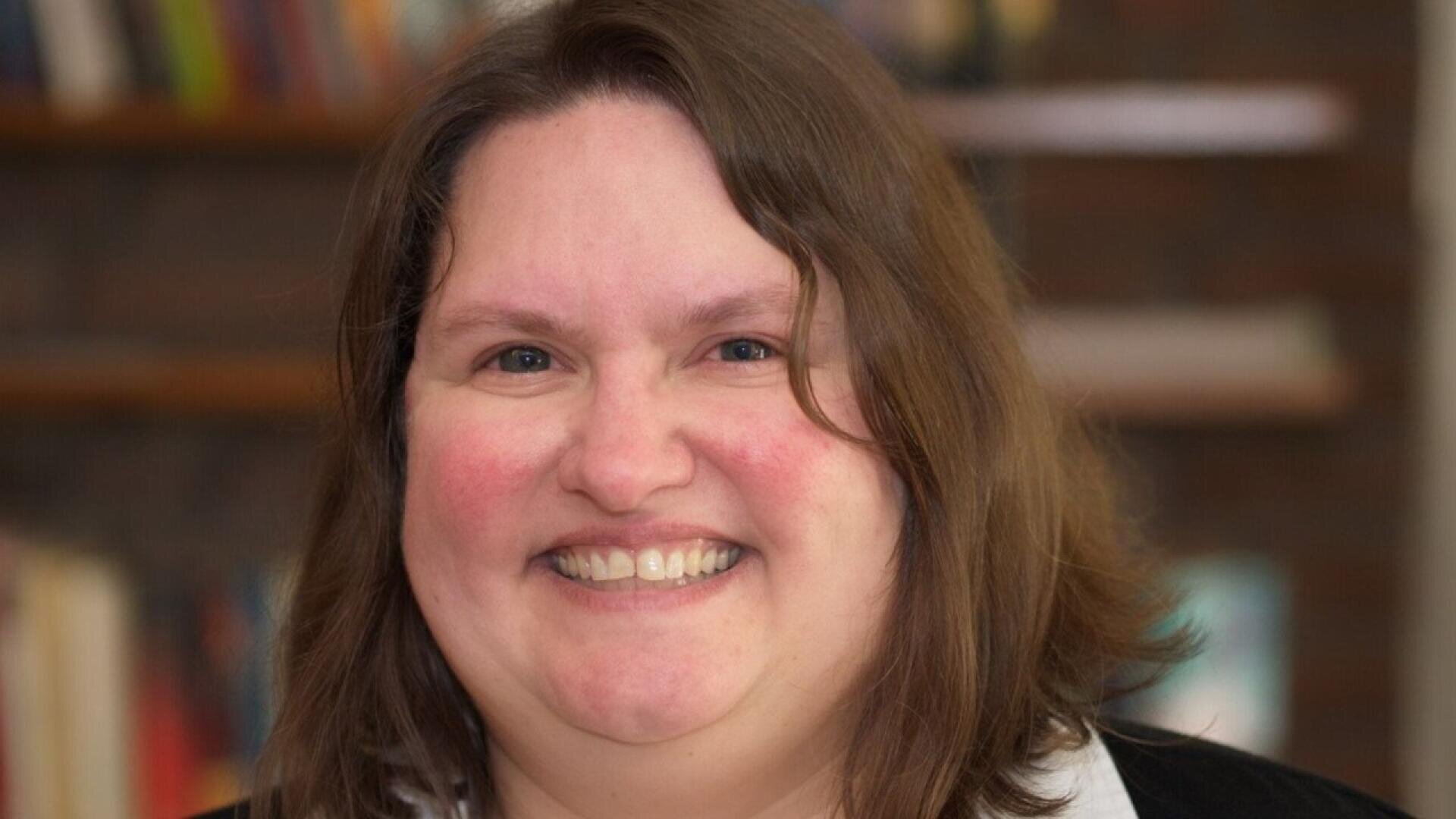 A woman with shoulder-length brown hair smiles warmly at the camera. She is indoors, with a blurry background that appears to show bookshelves.