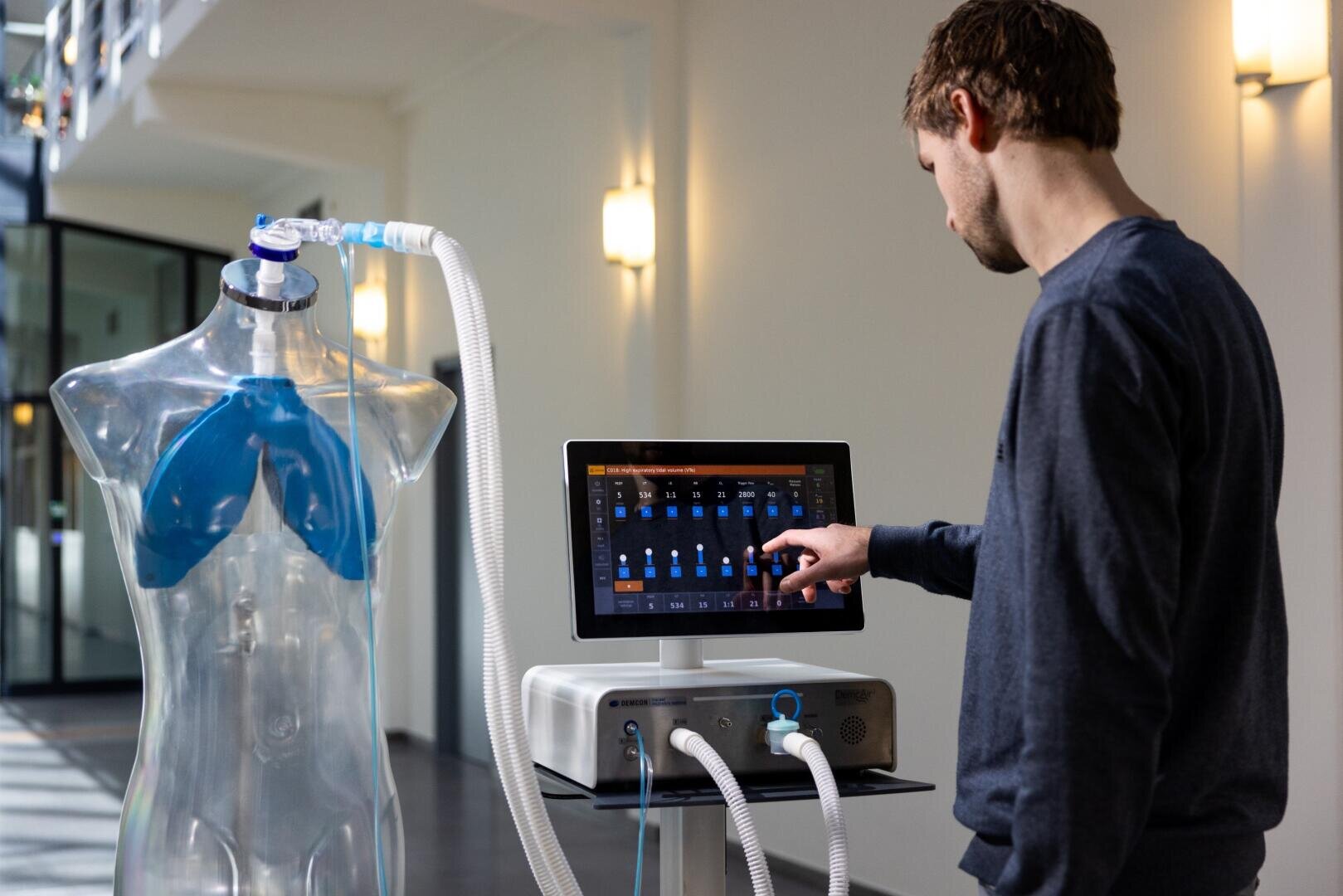A man adjusts settings on a touchscreen medical ventilator connected to a clear mannequin torso with visible blue lungs in a bright, modern indoor space.
