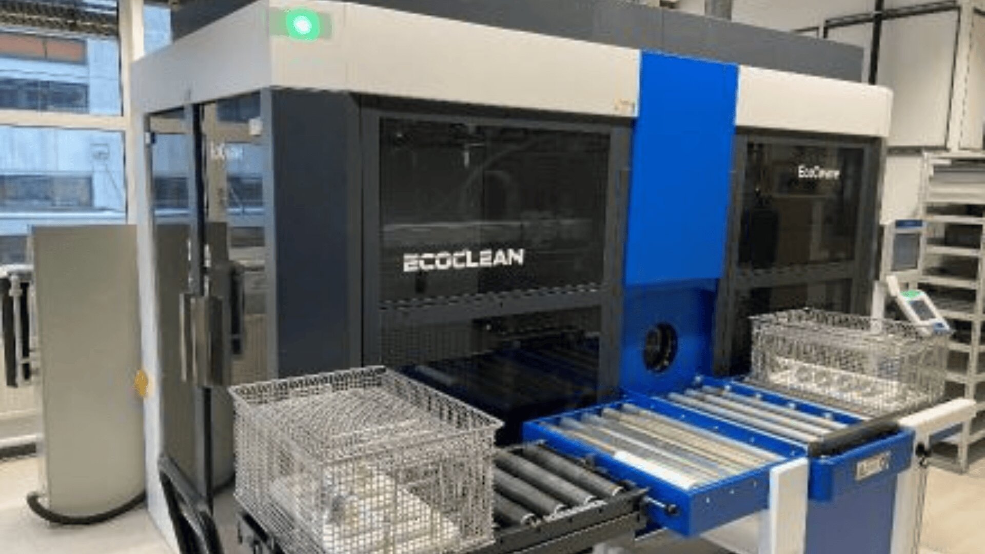 Eine große industrielle Ecoclean-Maschine mit zwei Metalldrahtkörben auf Rollen steht in einem sauberen, gut beleuchteten Raum mit Fenstern und Regalen im Hintergrund.