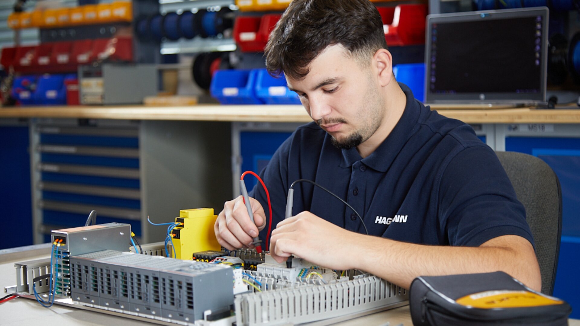 Ein Mann in einem marineblauen Poloshirt arbeitet in einem Elektroniklabor mit Sonden an der Verkabelung im Inneren eines offenen elektronischen Geräts. Im Hintergrund sind Werkzeuge und Geräte zu sehen.