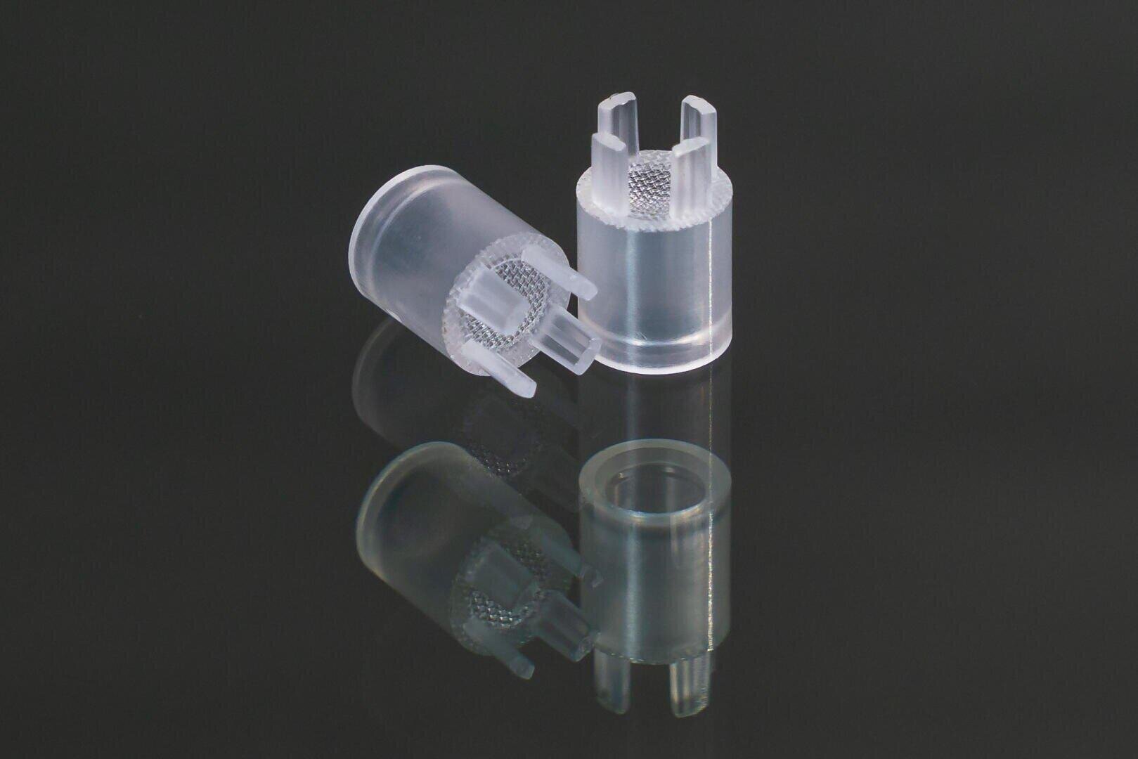 Two small, transparent, cylindrical plastic pieces with prongs and textured handles are depicted on a dark, reflective surface, with one lying on its side and the other standing upright.