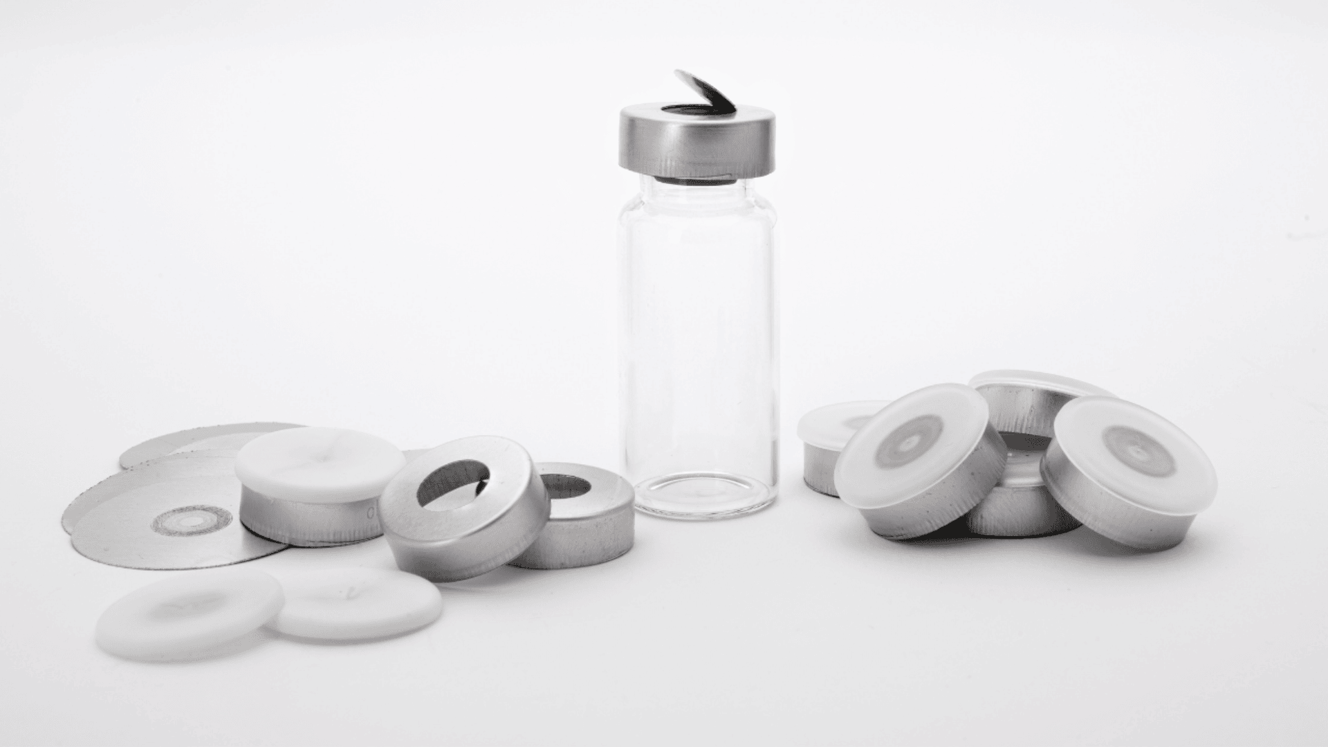 Ein Klarglasfläschchen mit einer teilweise aufgesetzten Metallkappe steht aufrecht, umgeben von mehreren losen Metallkappen und weißen Siegeln auf weißem Hintergrund.