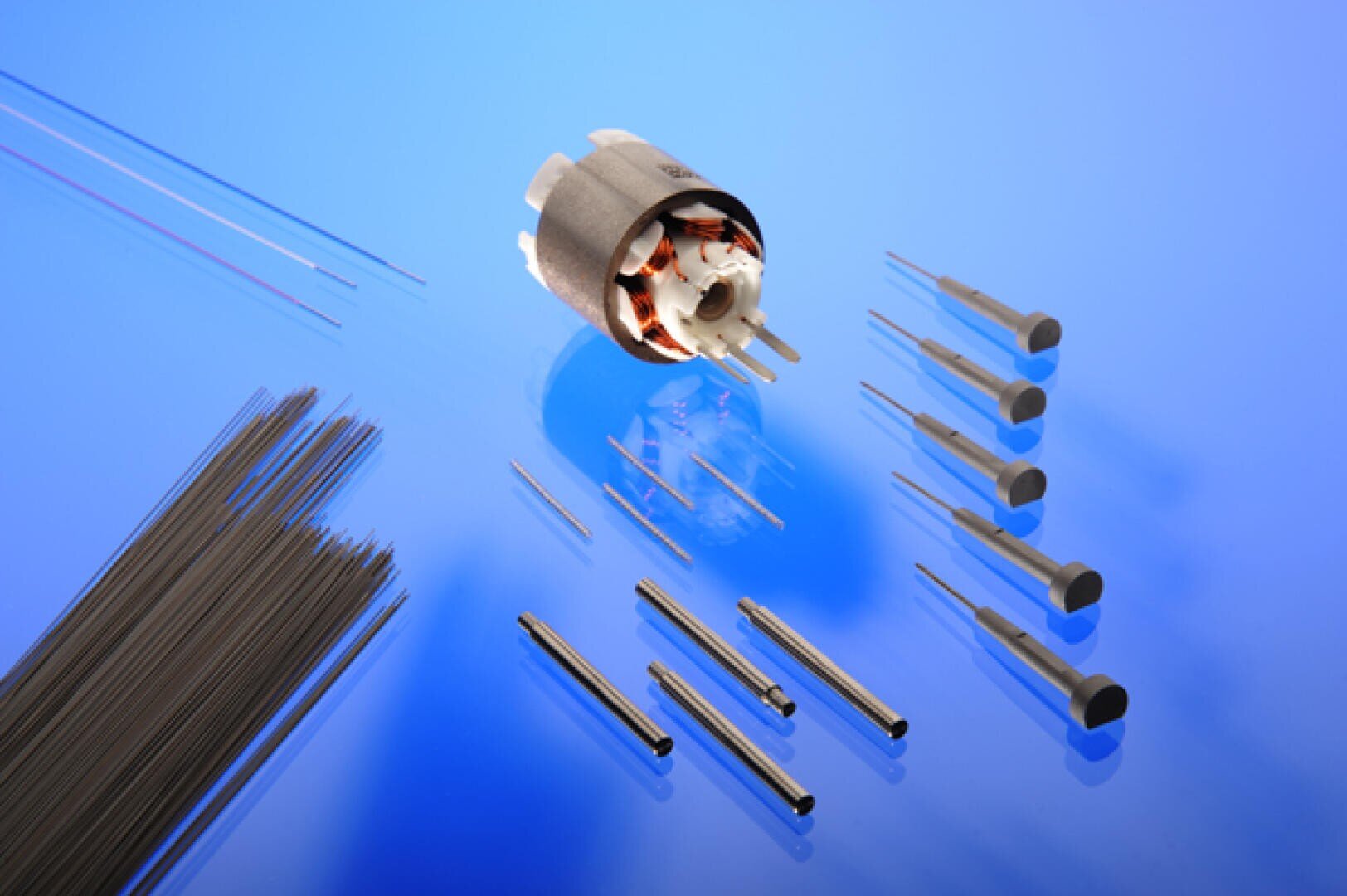 A variety of precision metal rods, wires, pins, and cylindrical electronic components are arranged on a reflective blue surface, with a small electric motor positioned near the center.
