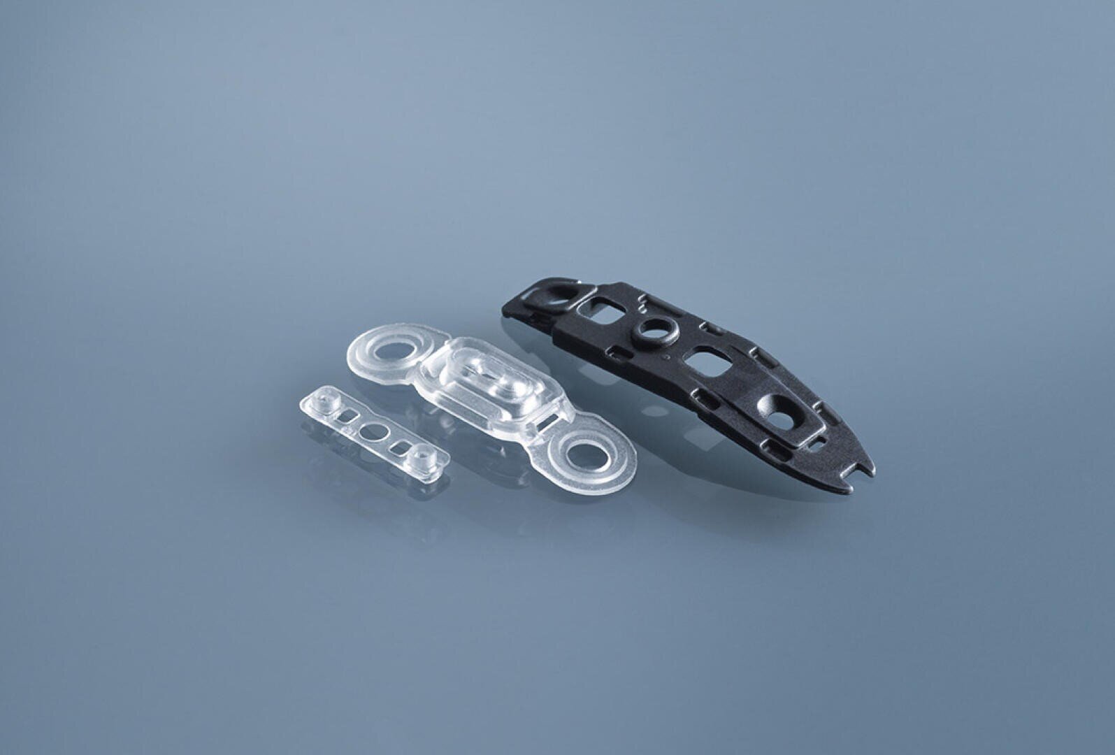 Three small plastic parts, including two clear parts and one black part, are shown on a smooth, gray-blue surface. The parts have rounded edges and holes, indicating that they are connectors or mechanical parts.
