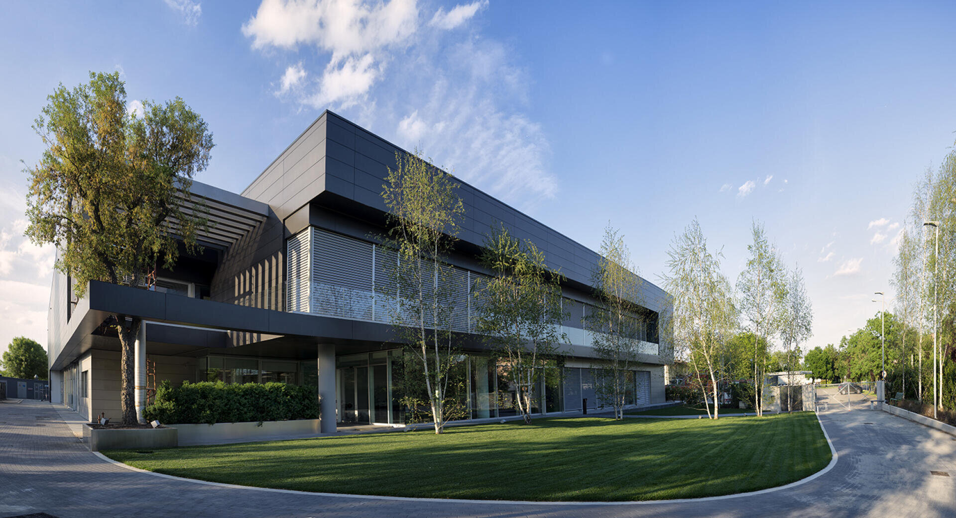 Ein modernes, zweistöckiges Gebäude mit dunkler, glatter Außenverkleidung, großen Fenstern und einem gepflegten Rasen, umgeben von jungen Bäumen unter einem blauen Himmel mit vereinzelten Wolken.
