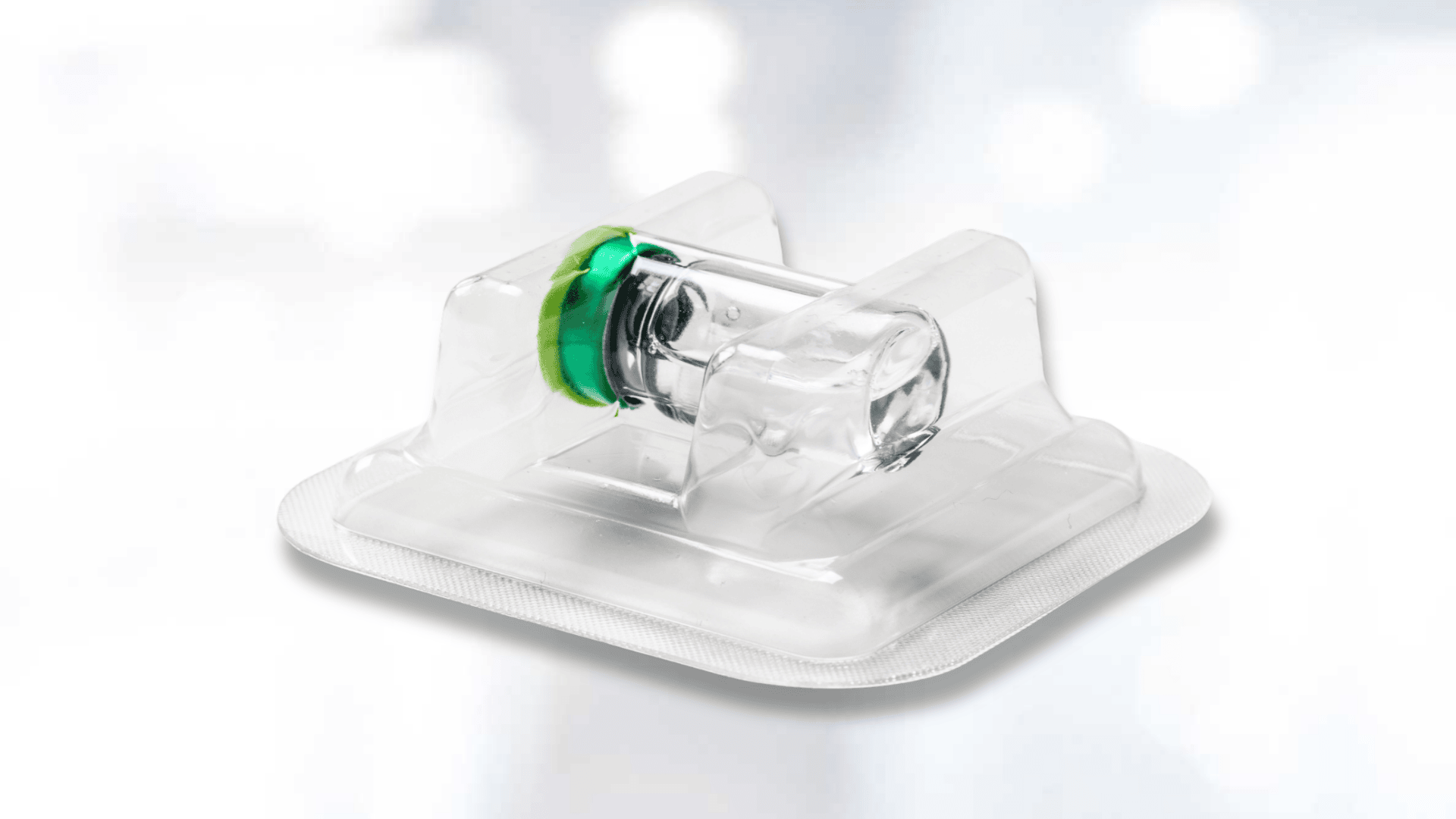 A clear glass vial with a green and black rubber stopper, sealed in transparent plastic packaging, placed on a white surface with a blurred light background.