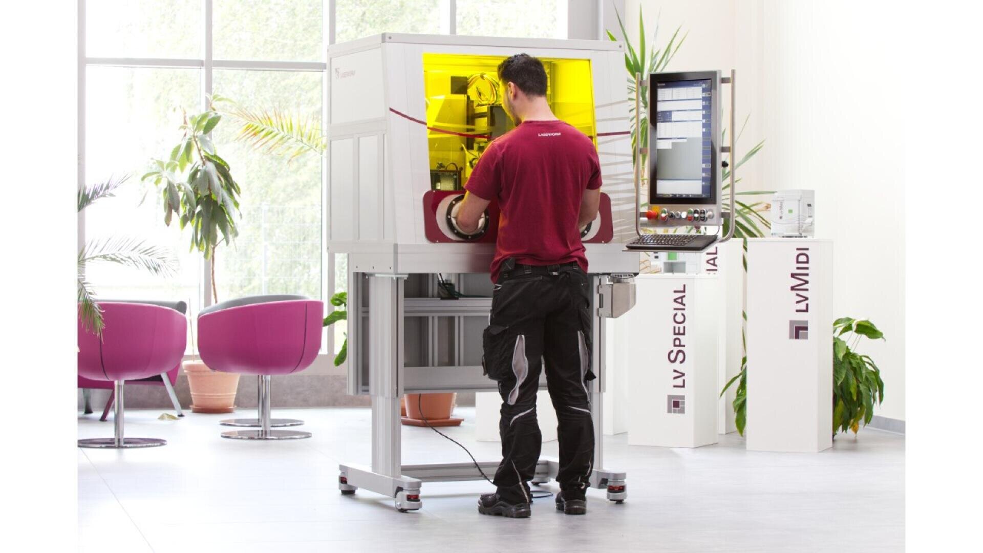 A man in a red shirt operates a machine with a yellow-lit chamber using built-in gloves, in a bright, modern room with large windows, plants, and magenta chairs.