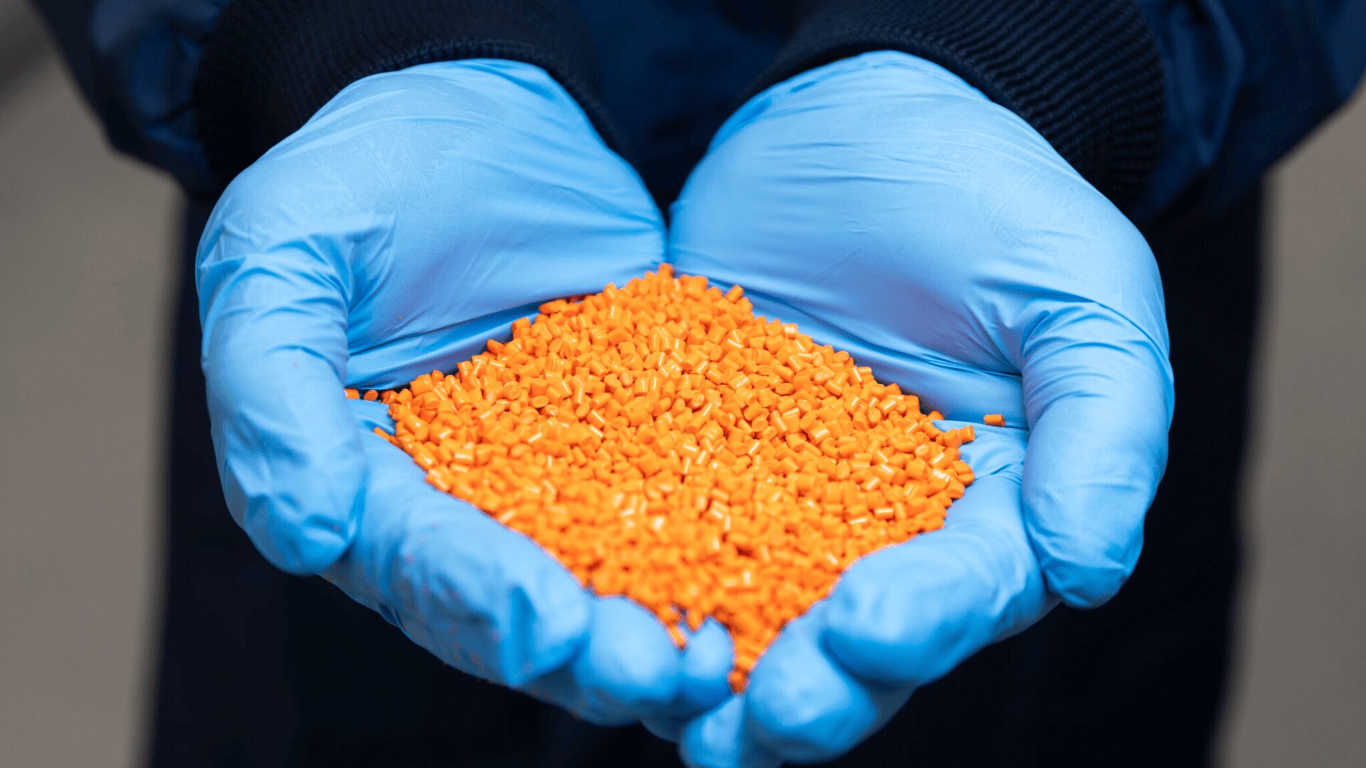 A person wearing blue gloves holds a pile of small, orange plastic pellets in their cupped hands.