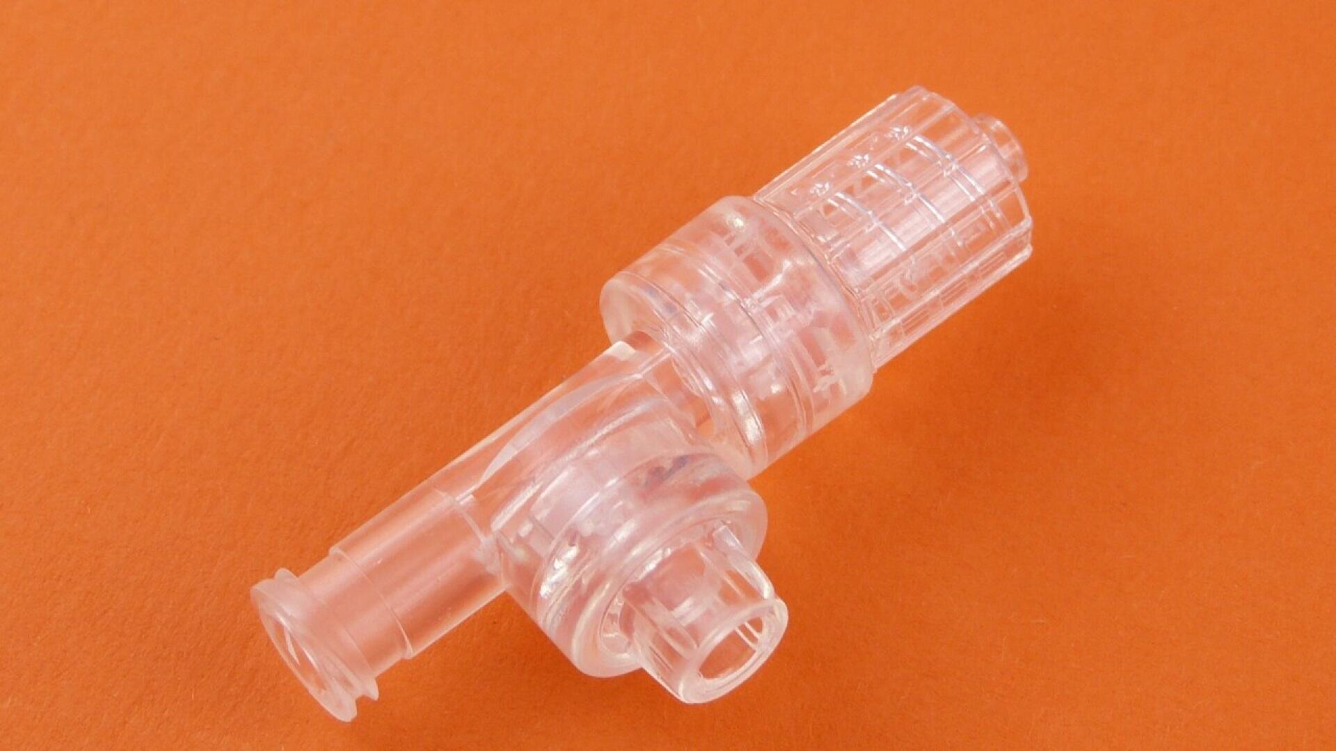 A transparent plastic medical connector with a cylindrical shape and textured grip, shown on an orange background.