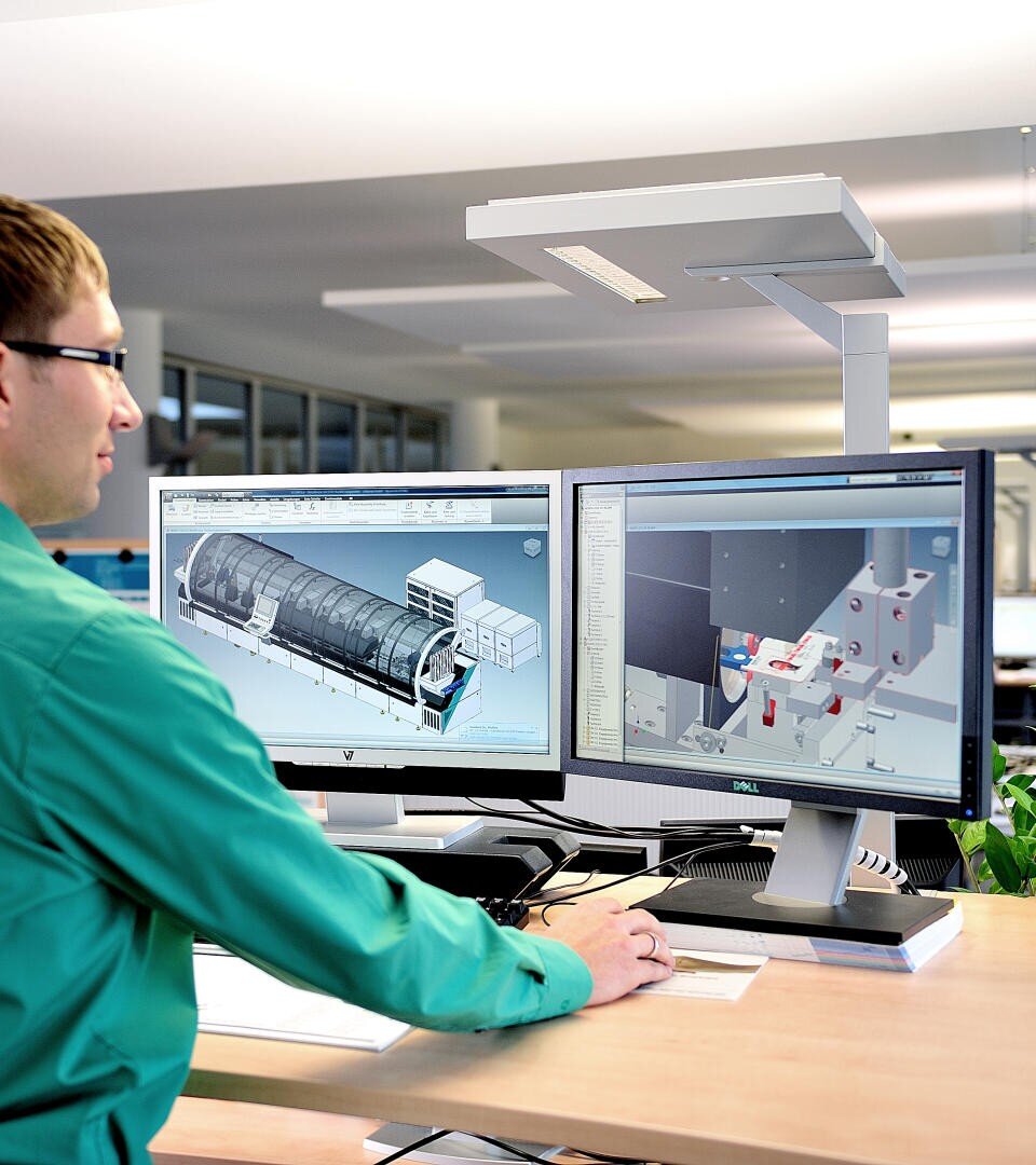 Eine Person in einem grünen Hemd arbeitet in einer modernen Büroumgebung an einem Schreibtisch mit zwei Computermonitoren, auf denen detaillierte 3D-Konstruktionsmodelle und Diagramme angezeigt werden.