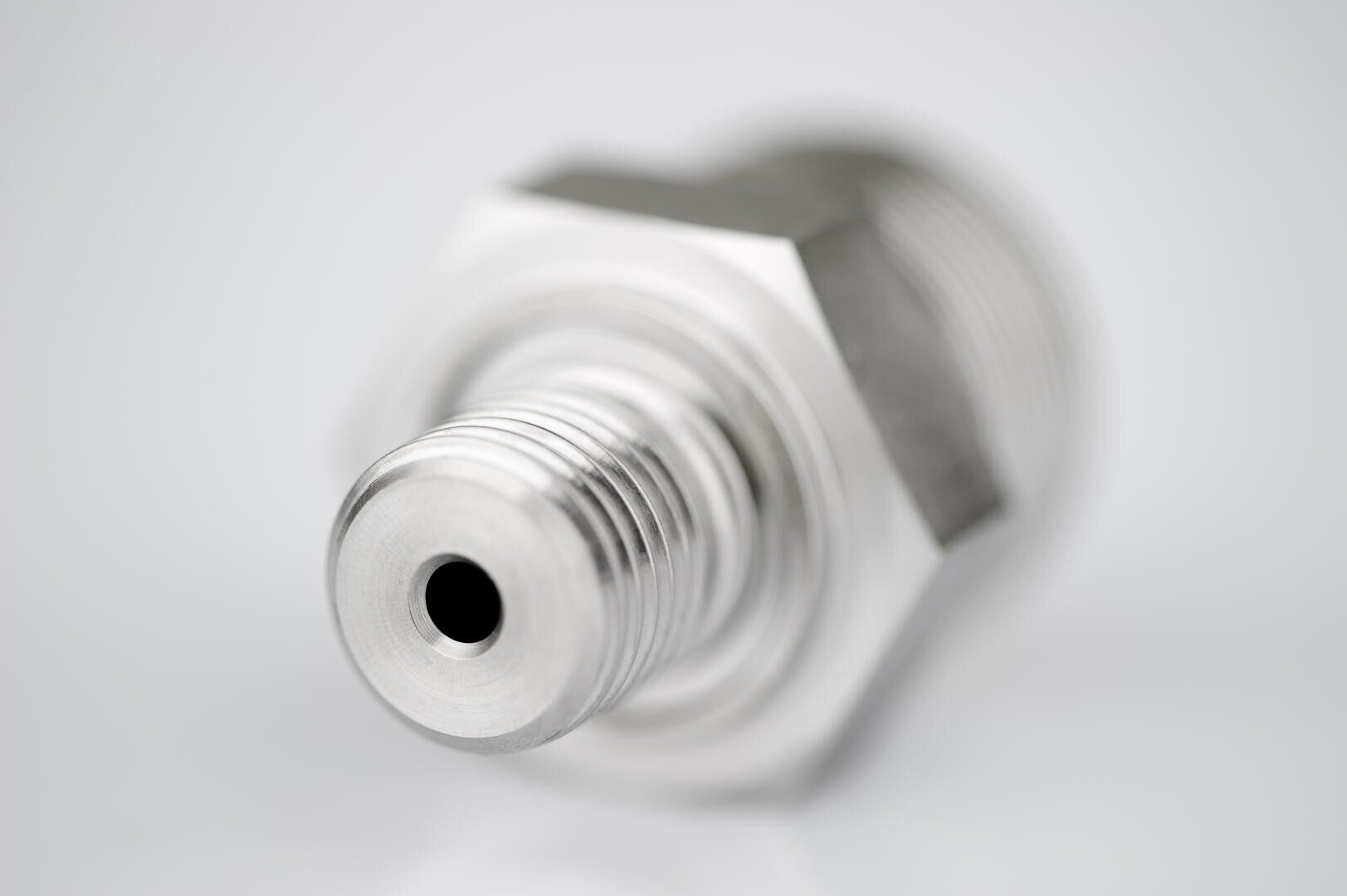 Close-up of a metal threaded connector or fitting, likely used in plumbing or machinery, with a smooth circular opening and hexagonal nut, set against a blurred, light gray background.