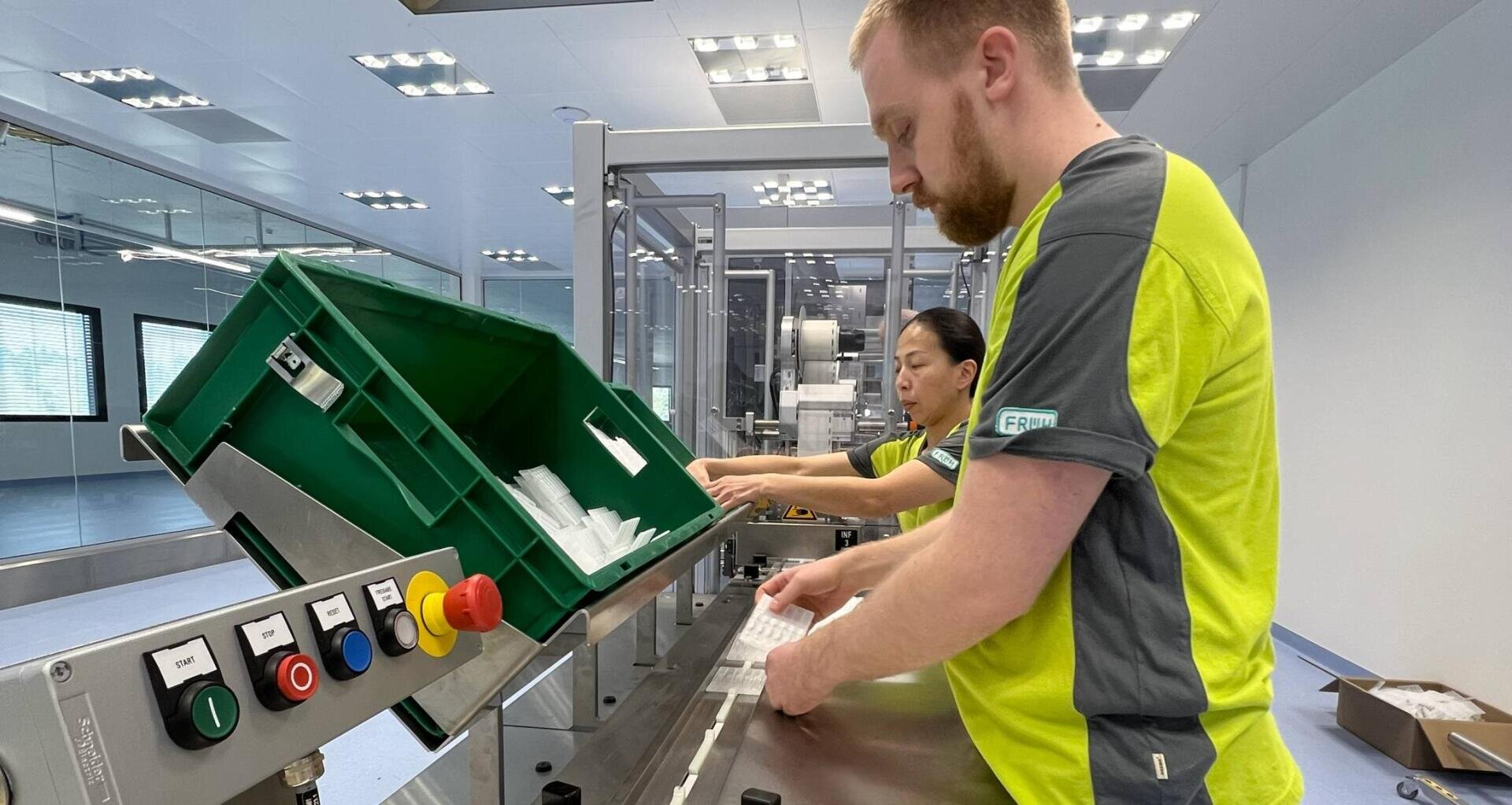 Zwei Arbeiter in einer sauberen, hell erleuchteten Fabrik sortieren oder prüfen Gegenstände aus einem großen grünen Behälter auf einem Förderband, umgeben von Industriemaschinen und Kontrollknöpfen.