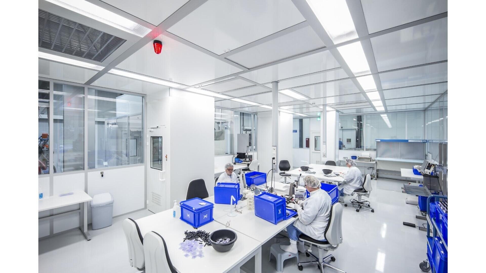 A modern, bright laboratory with people in cleanroom suits working at white tables, handling blue storage bins and scientific equipment. The lab is spacious and organized, with glass walls and overhead fluorescent lighting.