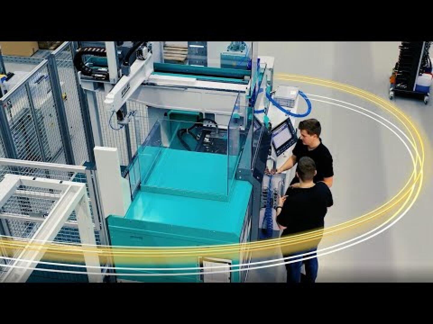 Two people operate a large, blue-green industrial machine in a factory environment surrounded by a metal fence; yellow and white swoosh graphics overlay the image, suggesting technology or automation.