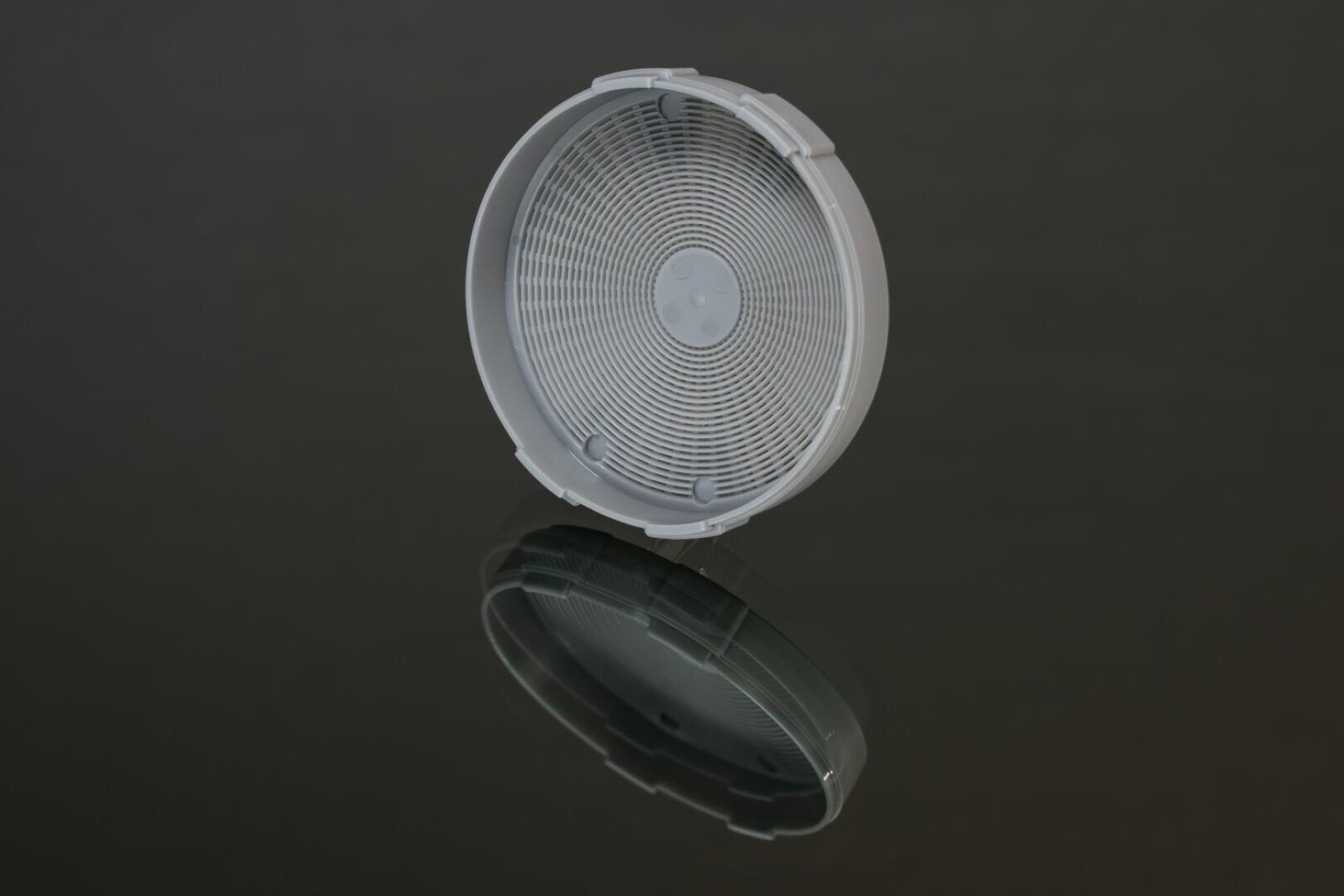 Ein runder, grauer Kunststoffgegenstand mit einem konzentrischen Kreismuster, möglicherweise eine Entlüftungsöffnung oder ein Deckel, wird auf eine spiegelnde, dunkle Oberfläche gelegt, so dass darunter eine deutliche Reflexion entsteht.