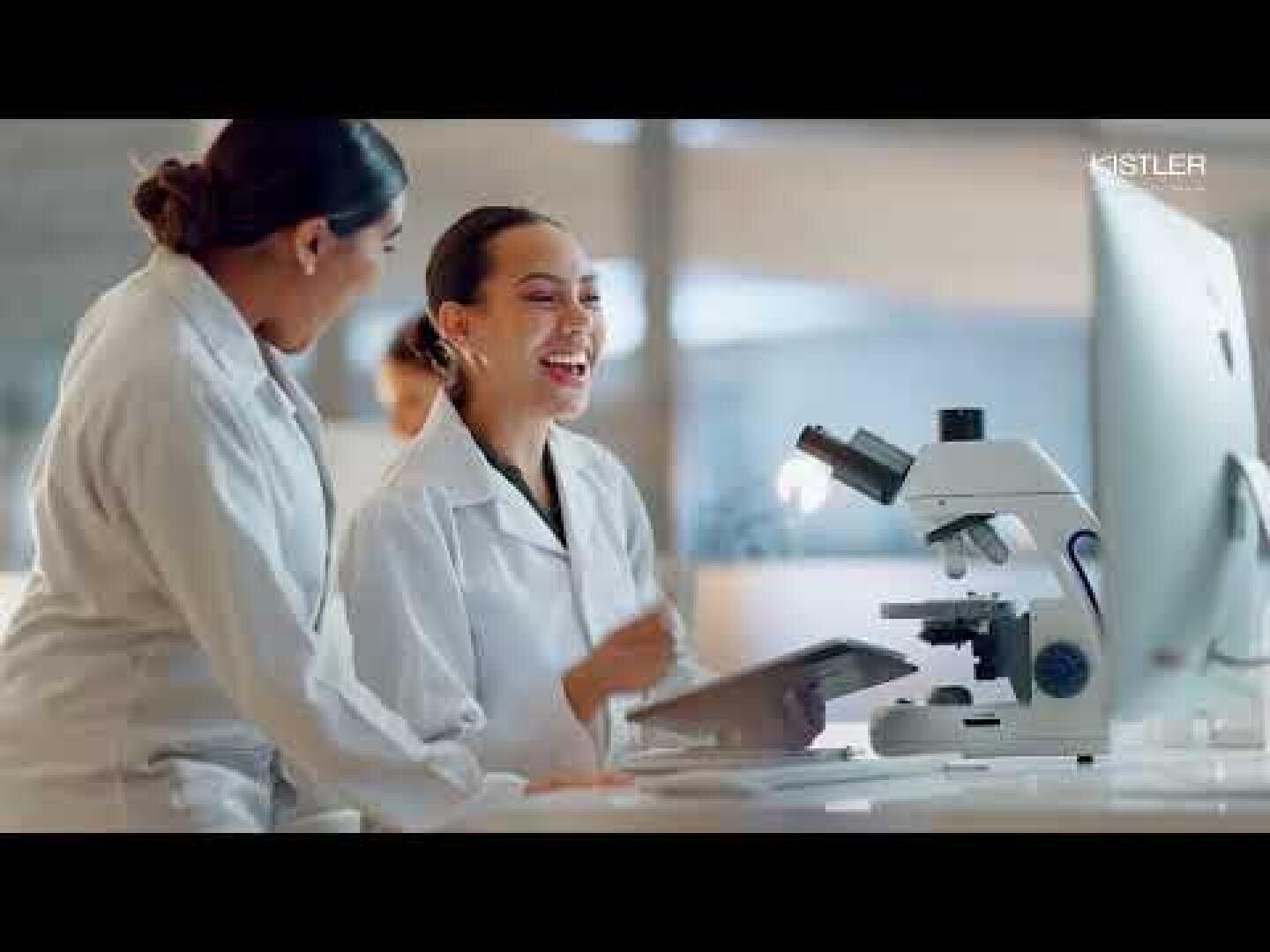 Zwei Wissenschaftlerinnen in weißen Laborkitteln lächeln und unterhalten sich, während sie in einem Labor an einem Computer und einem Mikroskop arbeiten.
